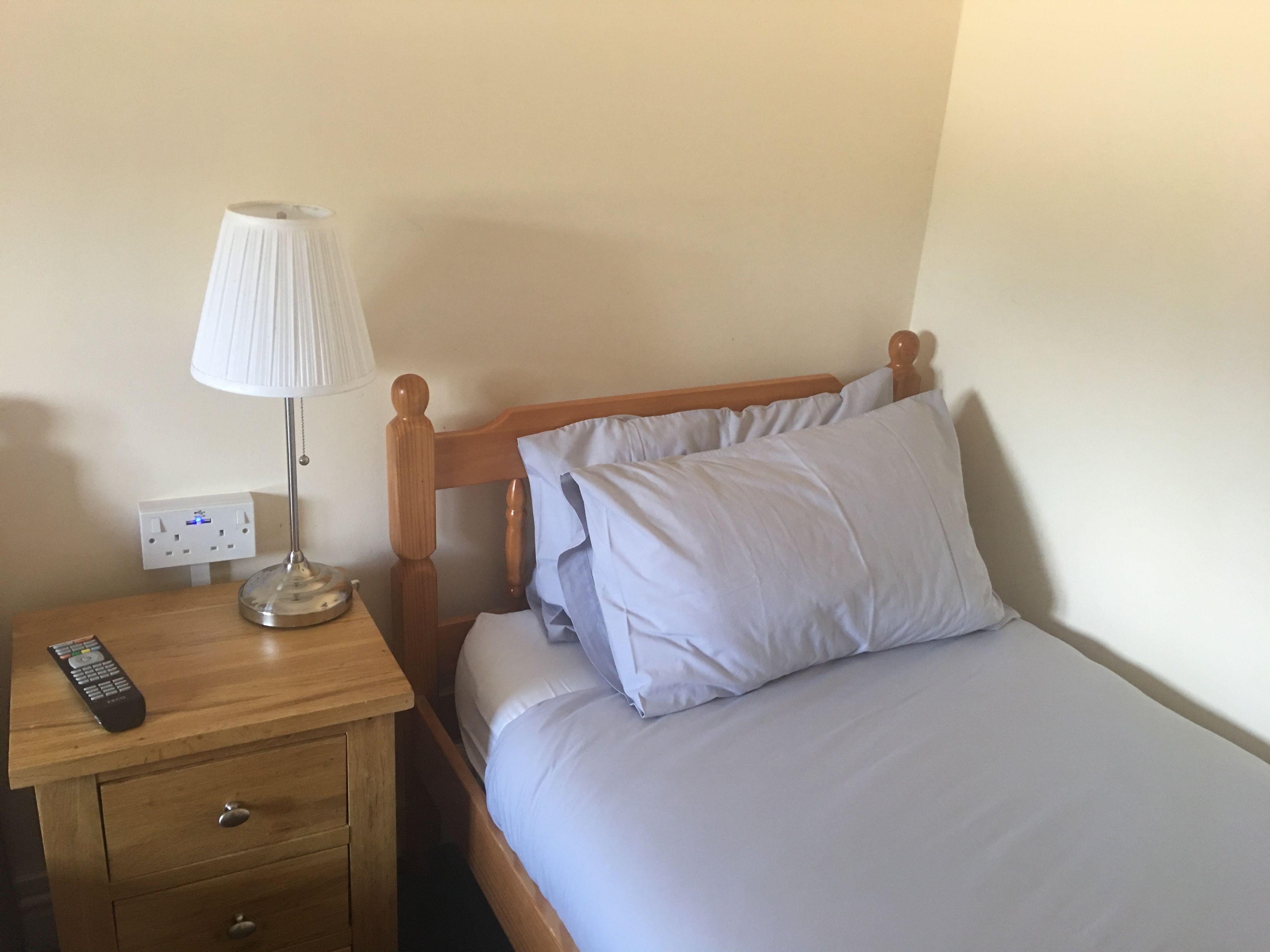 Cama bem arrumada com roupa de cama branca, mesa de cabeceira de madeira com abajur e controle remoto.