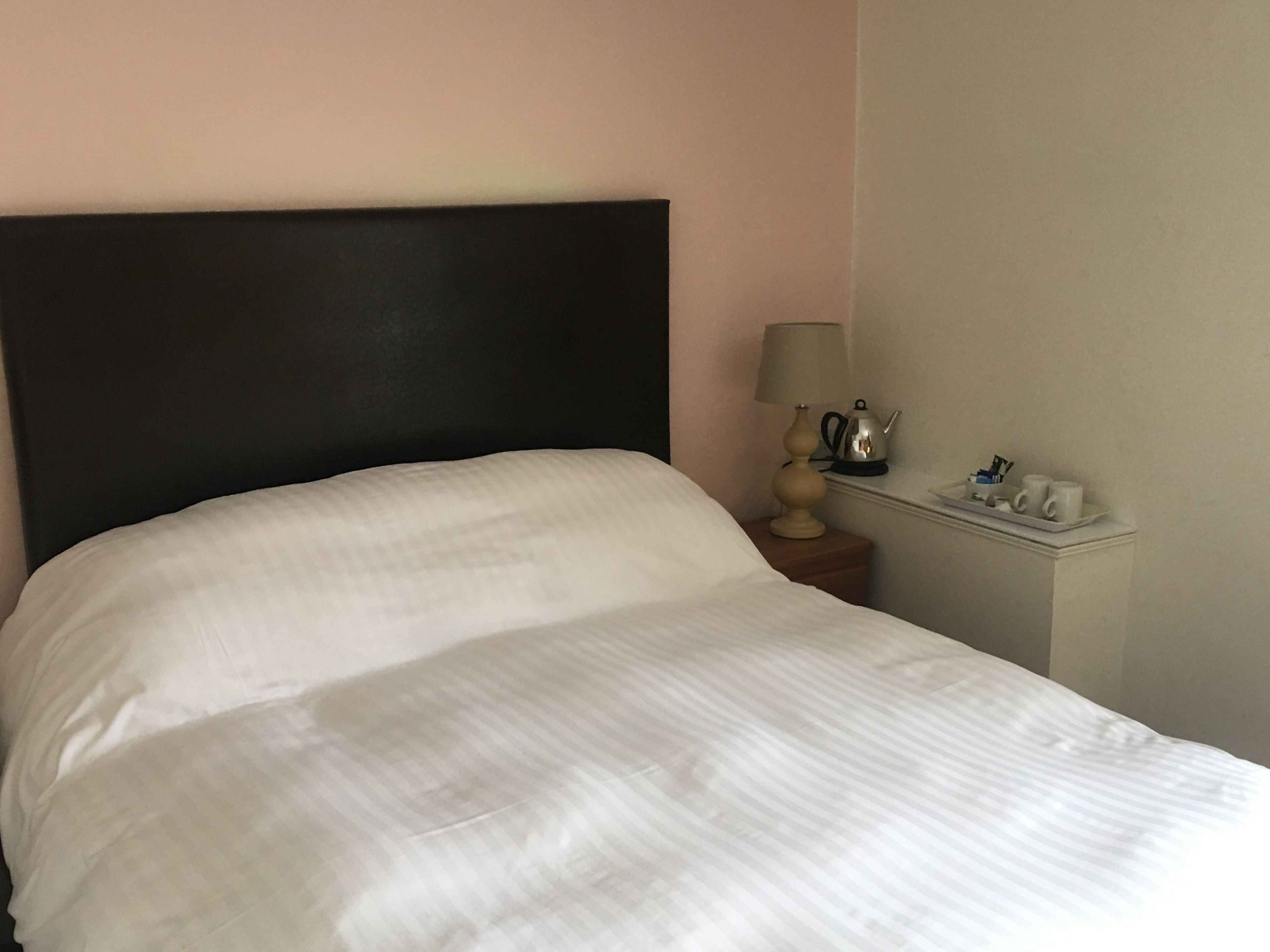 A neatly made bed with a dark headboard, a bedside table with a lamp, and a small tray with cups and a kettle.