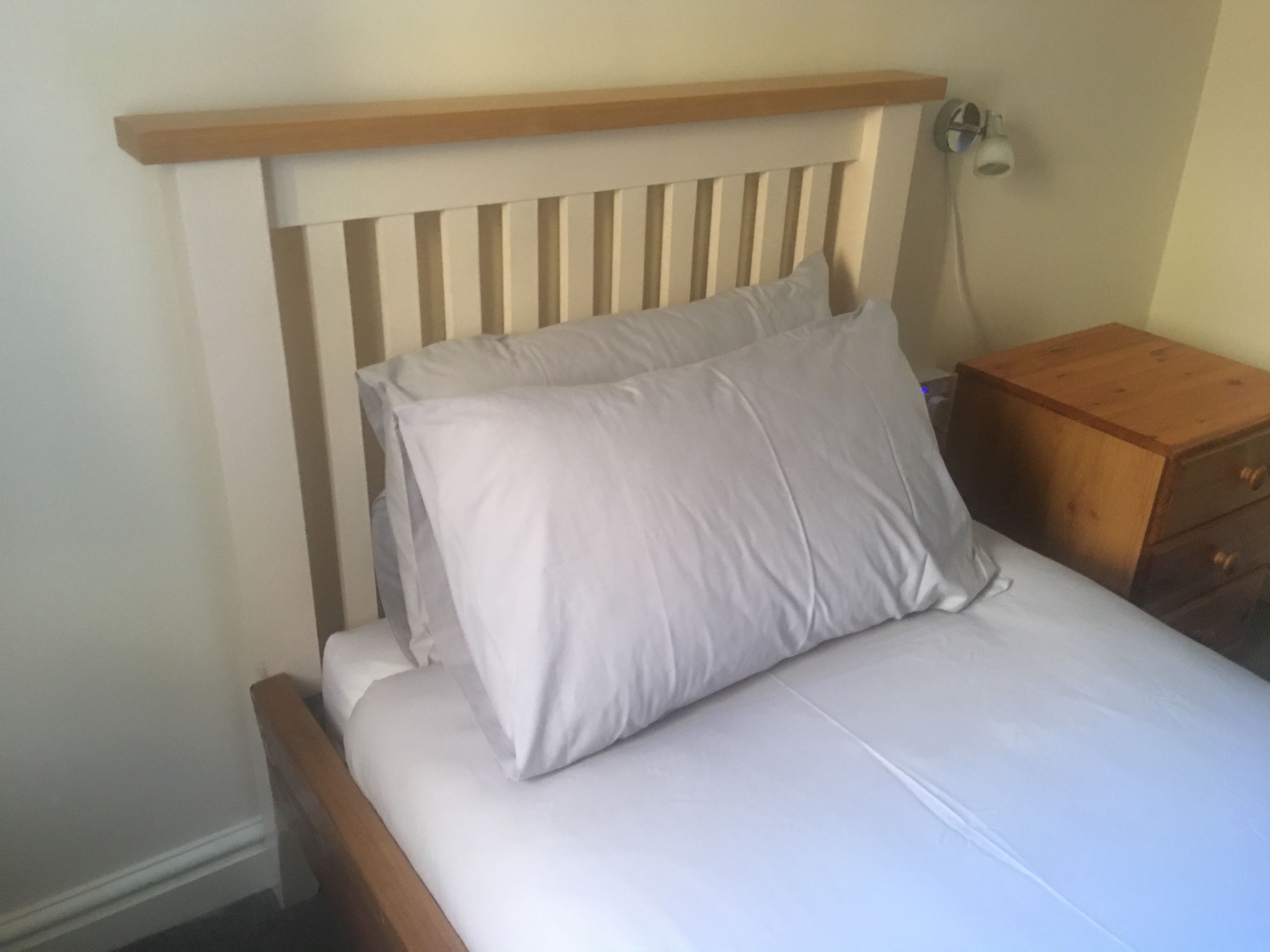 A bed with a wooden headboard and two pillows, a wooden nightstand with drawers, and a wall-mounted lamp.
