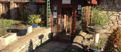 A stone building with a wooden entrance, potted plants, and a sign indicating reception contact numbers.