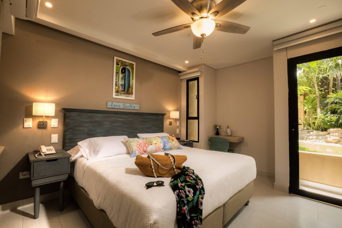 Une chambre à coucher avec un lit, des lampes de chevet, un ventilateur de plafond, une table de chevet, un téléphone, un coussin à motifs floraux, un sac et une fenêtre donnant sur un paysage verdoyant.
