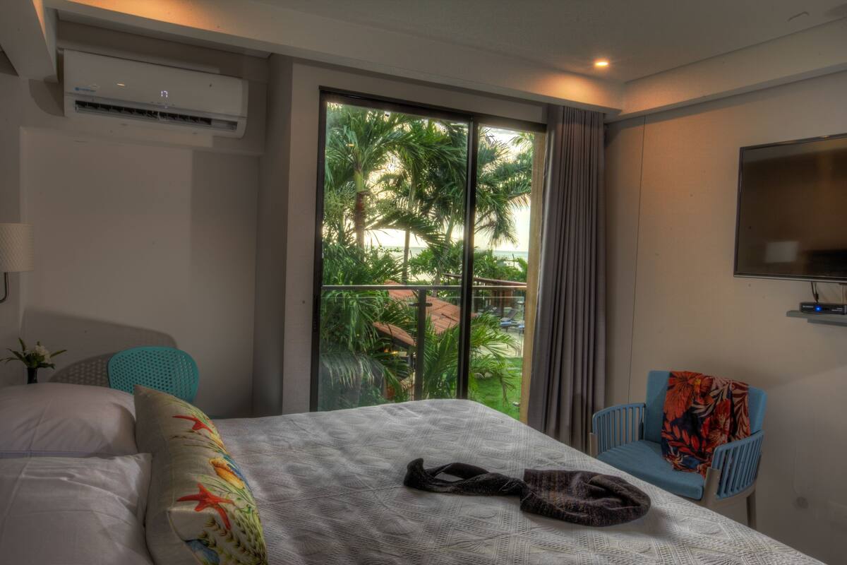 Une chambre d’hôtel avec un grand lit, un téléviseur fixé au mur et une vue sur les palmiers par la fenêtre.