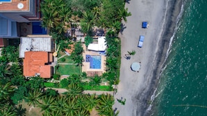 An aerial view of a coastal property with a swimming pool, a tennis court, and a beach area with umbrellas.