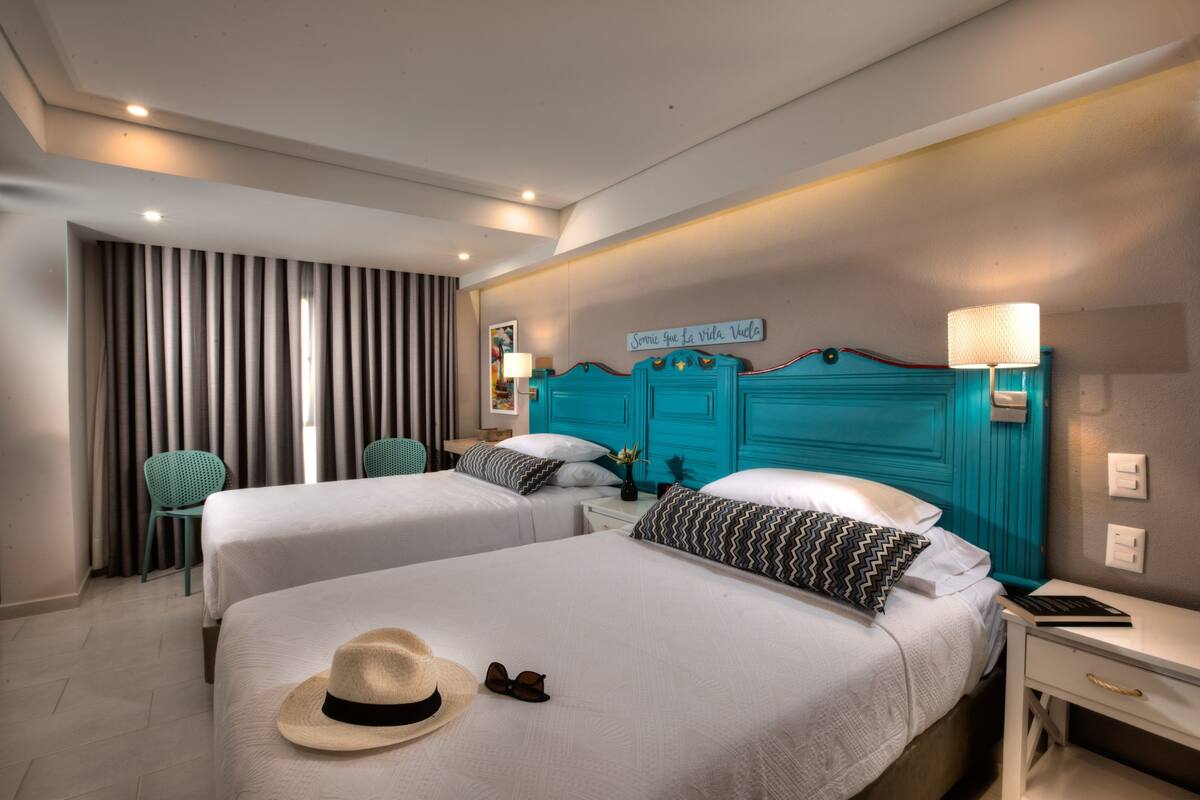 Une chambre d’hôtel avec deux lits, une tête de lit de couleur turquoise, un panneau décoratif au mur, une lampe et un chapeau posé sur l’un des lits.