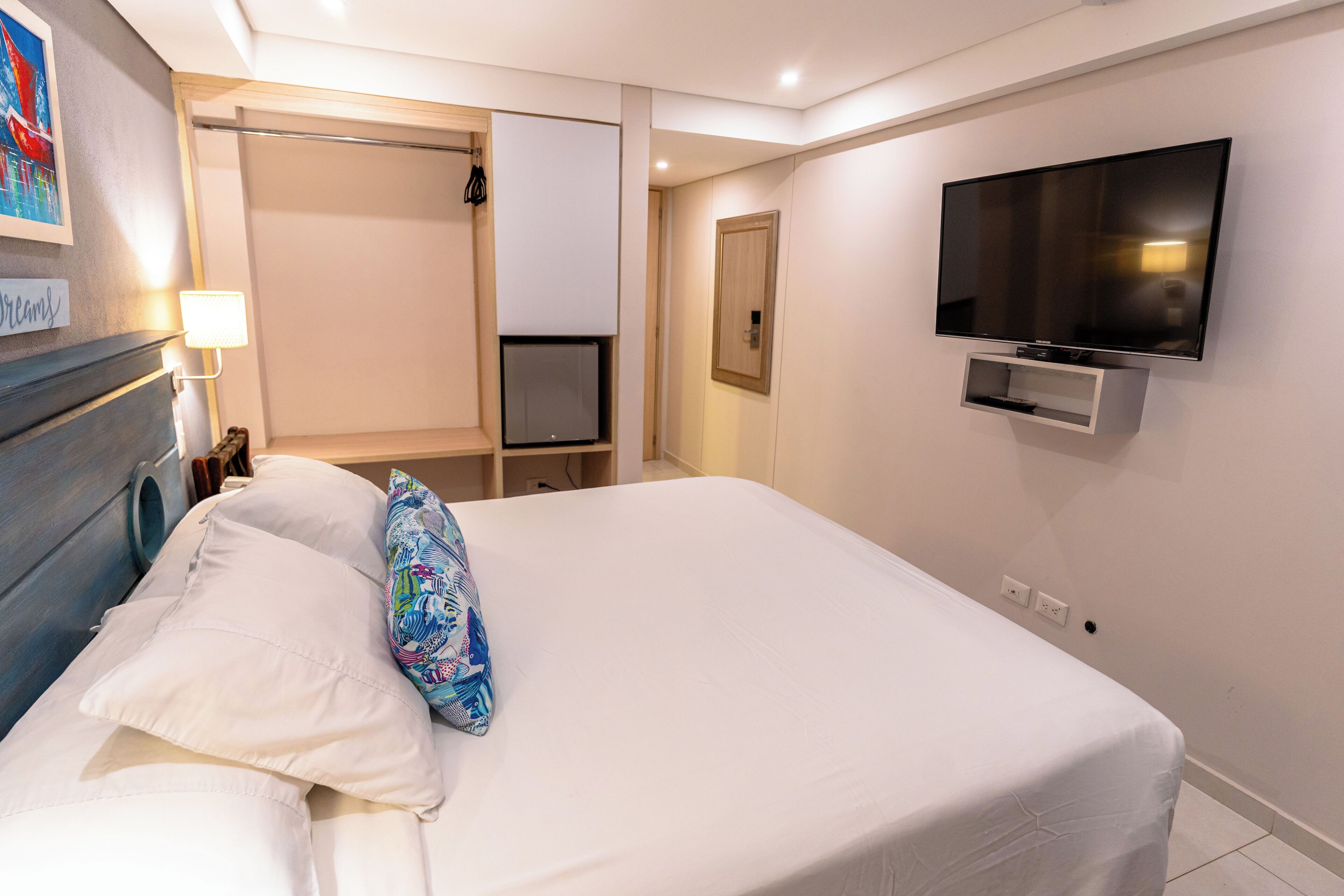 Habitación de hotel con cama, televisión montada en la pared, una puerta y un pequeño estante.