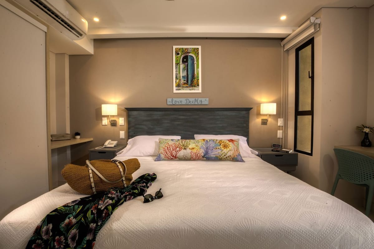 Un lit bien fait, agrémenté d’un coussin à motifs floraux, d’une lampe de chevet et d’une inscription au-dessus du lit qui dit : « Aimez profondément ».