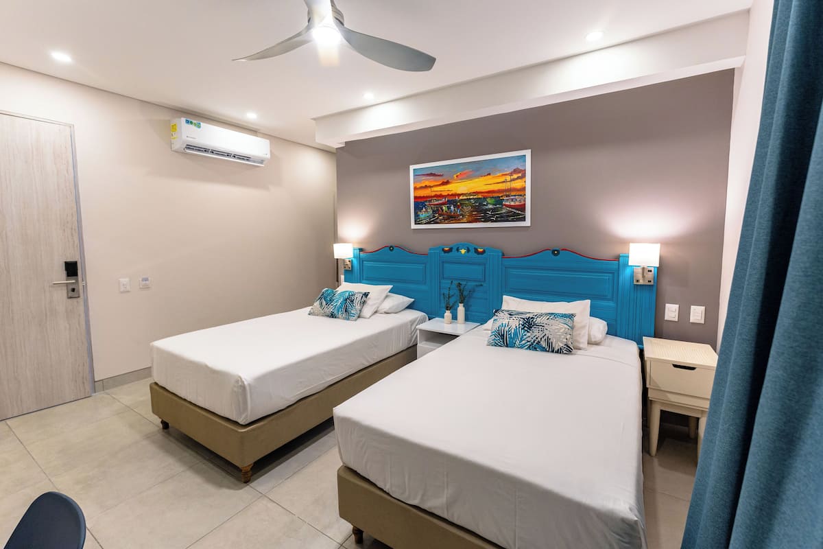Une chambre d’hôtel avec deux lits, un ventilateur de plafond, un climatiseur, un tableau au mur et une table de chevet avec une lampe.