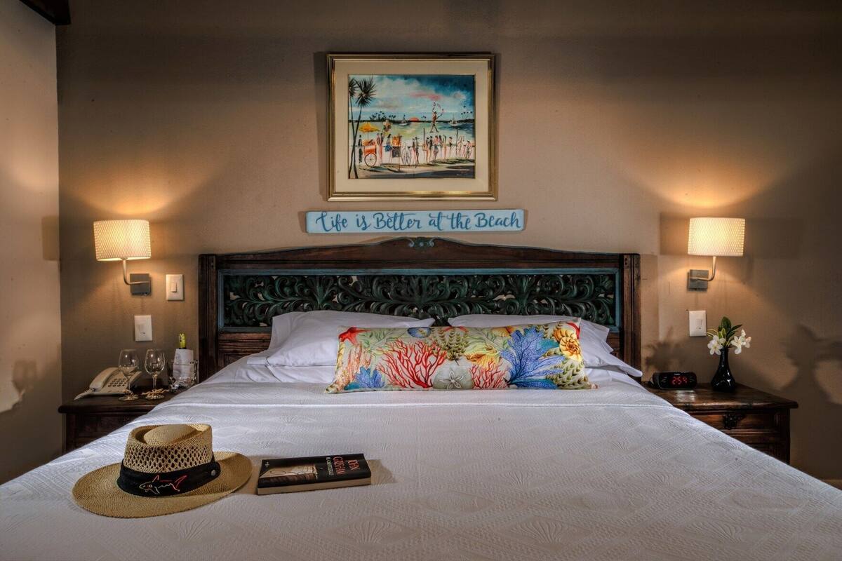 Ein Hotelzimmer mit einem Bett, einem Bild mit Strandmotiv, einem Schild mit der Aufschrift 'Life is Better at the Beach', zwei Nachttischlampen, einem Nachttisch mit Telefon und digitaler Uhr sowie einem Hut und einem Buch auf dem Bett.