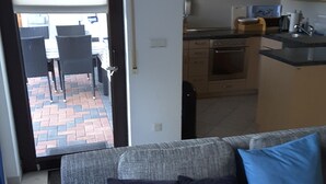 An interior with a sofa, a dining table, chairs, a kitchen area, and a view through a door to an outdoor patio. - Ferienwohnung - Zoe (Sankt Wendel)