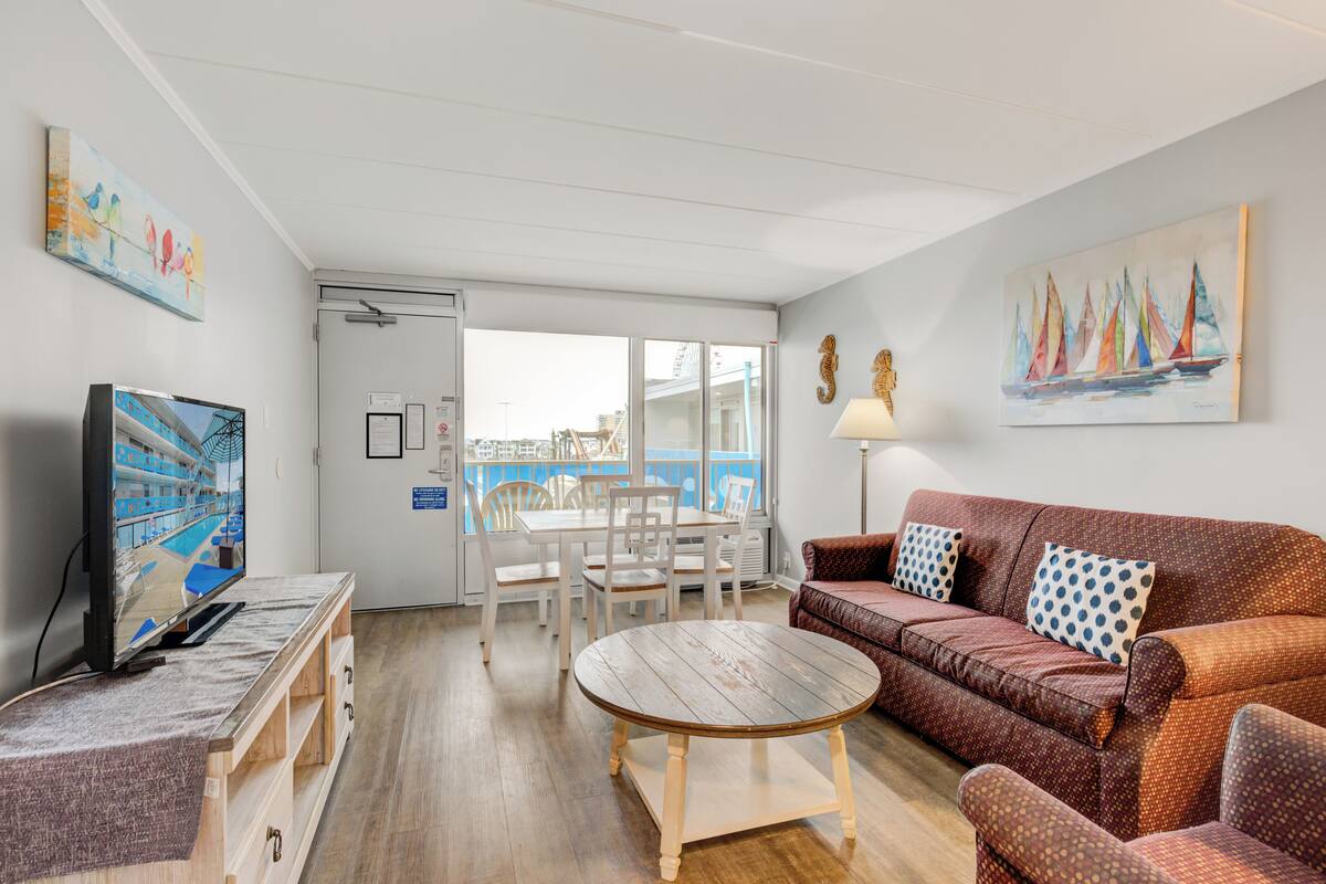 Una sala de estar moderna con una mesa de centro de madera, un sofá marrón con cojines estampados, un televisor sobre un mueble de madera, una mesa de comedor con sillas y un amplio ventanal con vistas a un edificio.