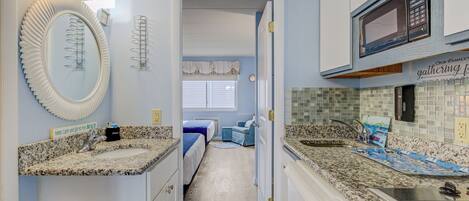 A compact kitchen with granite countertops, a microwave, and a sink. A hallway leads to a bedroom with a bed and a window.