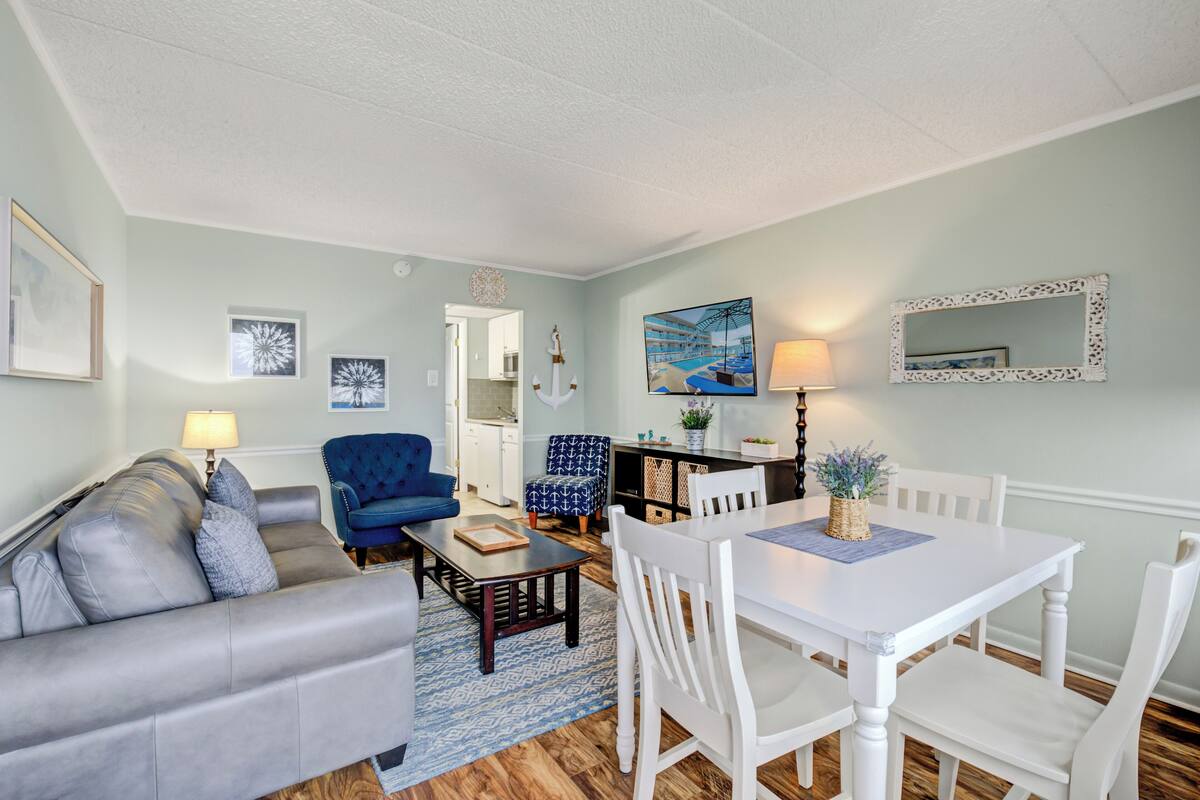 Una sala de estar con un sofá gris, un sillón azul, una mesa de comedor blanca con cuatro sillas y una zona de cocina al fondo.
