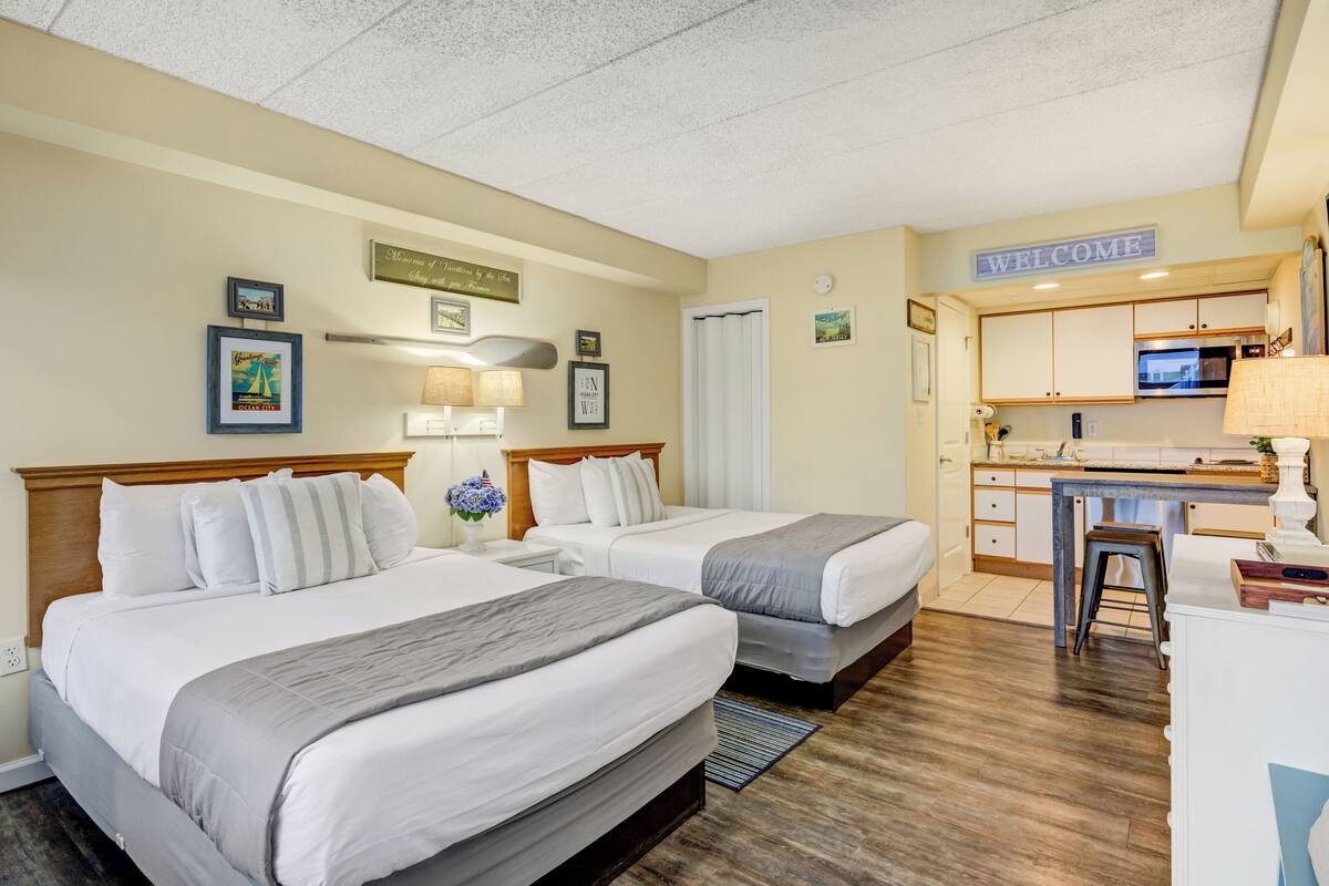 Habitación de hotel con dos camas, kitchenette, un cartel de 'Welcome', cuadros en la pared y un cartel que dice 'Monuments of the Sea'.
