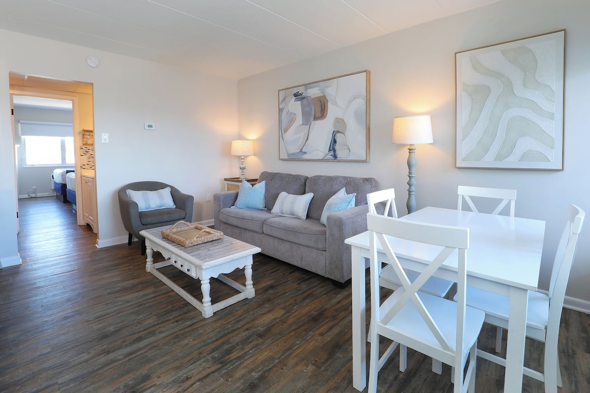 Una sala de estar con un sofá gris, una mesa de centro blanca y una mesa de comedor blanca con sillas. Hay dos lámparas fijadas a la pared y un cuadro abstracto grande sobre el sofá.