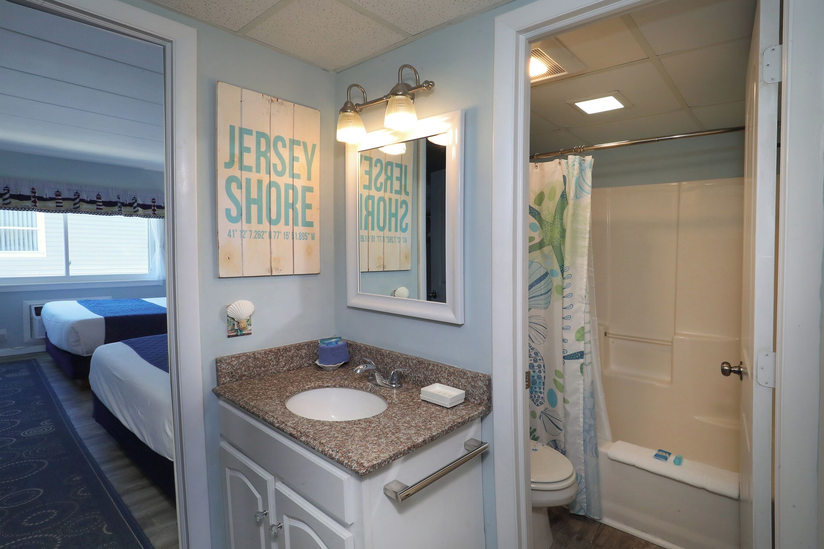 A hotel bathroom with a sink, mirror, shower, and two beds in the adjacent room.