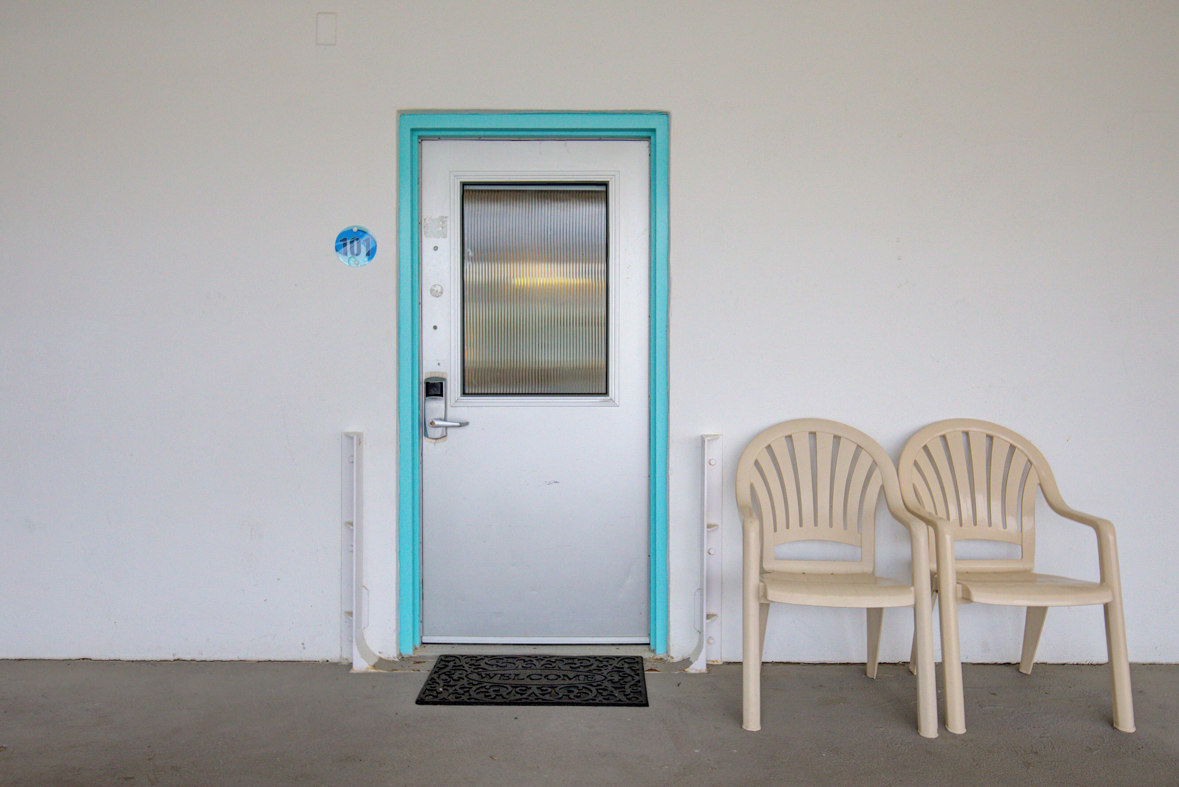 A white door with a turquoise frame, a small window, and a doorbell. There are two beige plastic chairs next to the door.