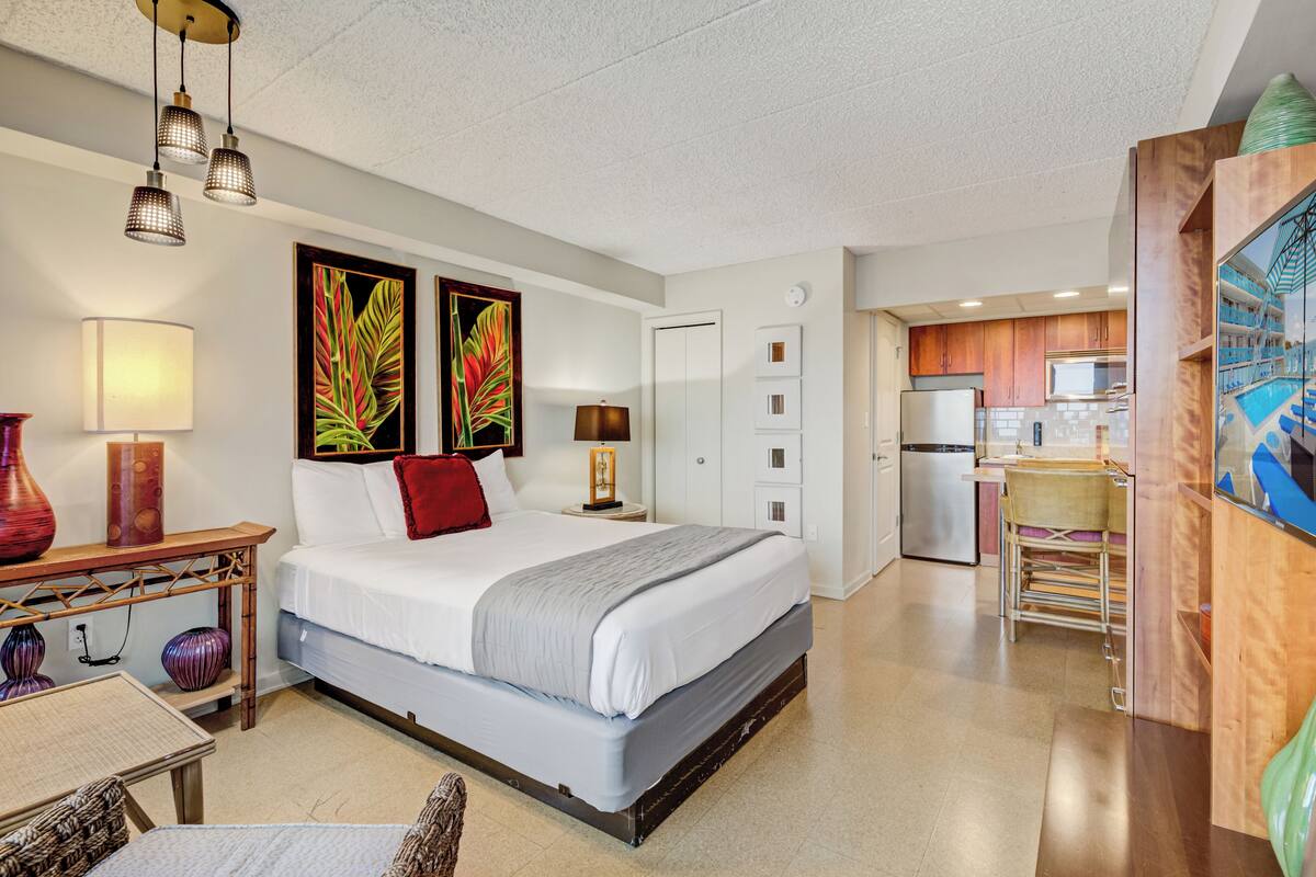 Habitación de hotel con cama, mesita de noche, lámpara y obra de arte. Cuenta con una kitchenette equipada con refrigerador y microondas.