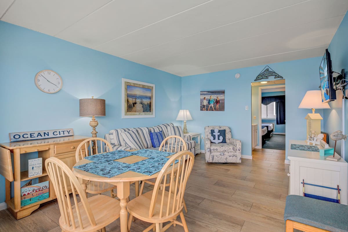 Una habitación con una mesa de comedor, sillas, un sofá y un televisor. Hay un cartel que dice "Ocean City" y una fotografía de una playa.