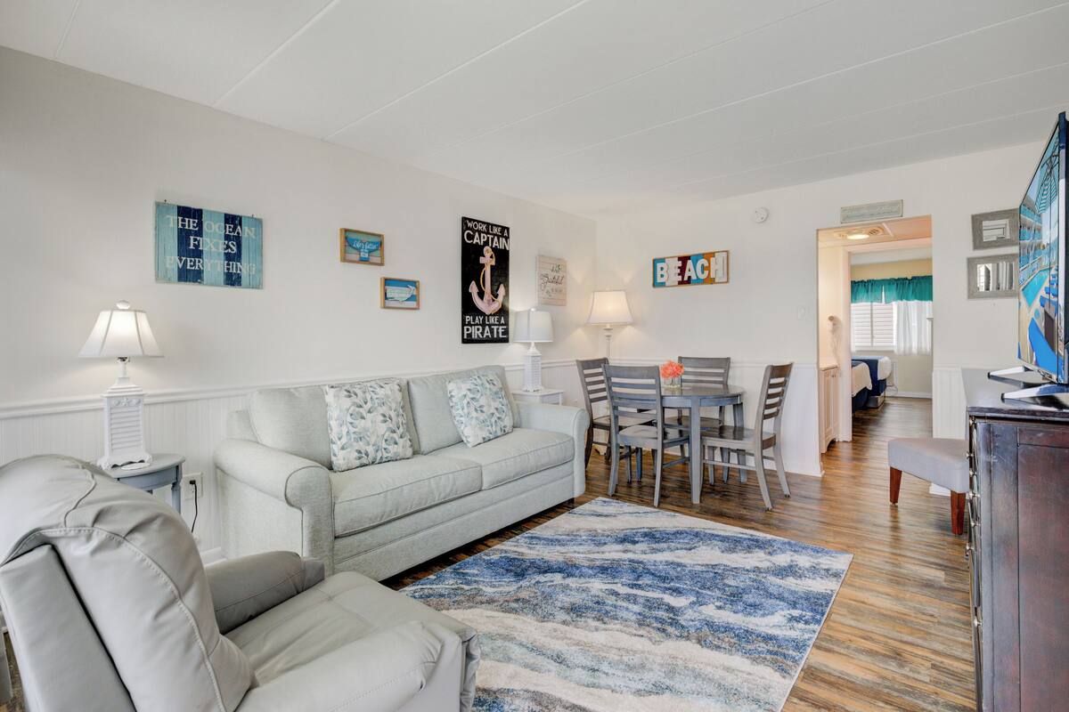 Una sala de estar con un sofá de gris claro, un tapete azul y blanco, y un comedor con una mesa y sillas.