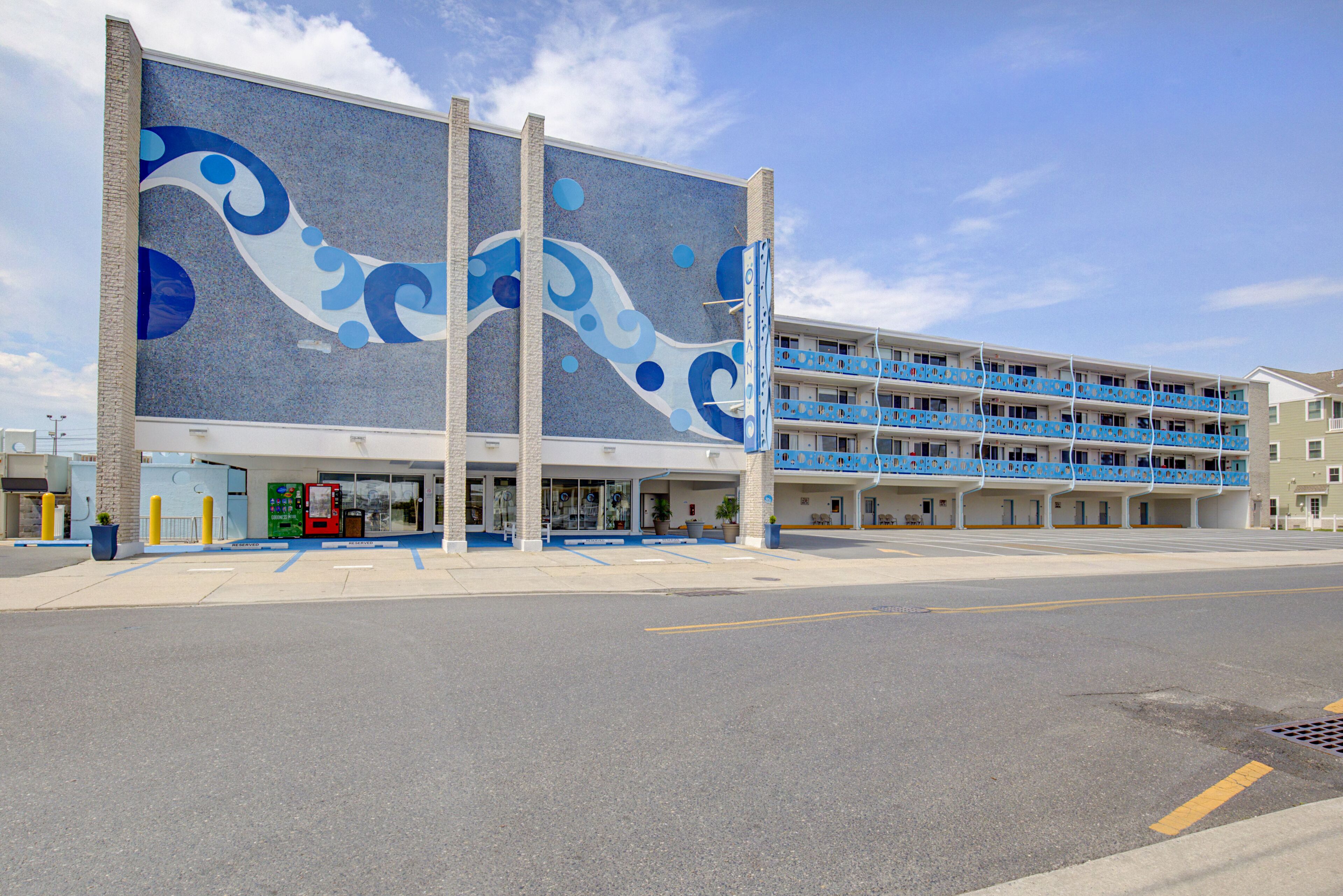 Un hôtel à plusieurs étages, dont la façade est ornée d’une fresque remarquable aux couleurs bleu et blanc, avec un parking et un ciel dégagé.