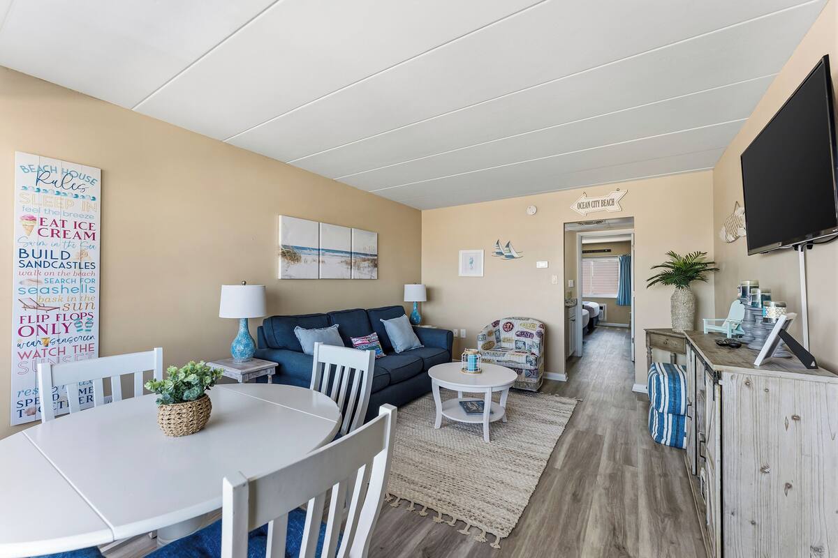Una sala de estar acogedora con un sofá, una mesa de centro y una mesa de comedor. Hay un televisor montado en la pared y un letrero que dice 'BEACH HOUSE RULES'.