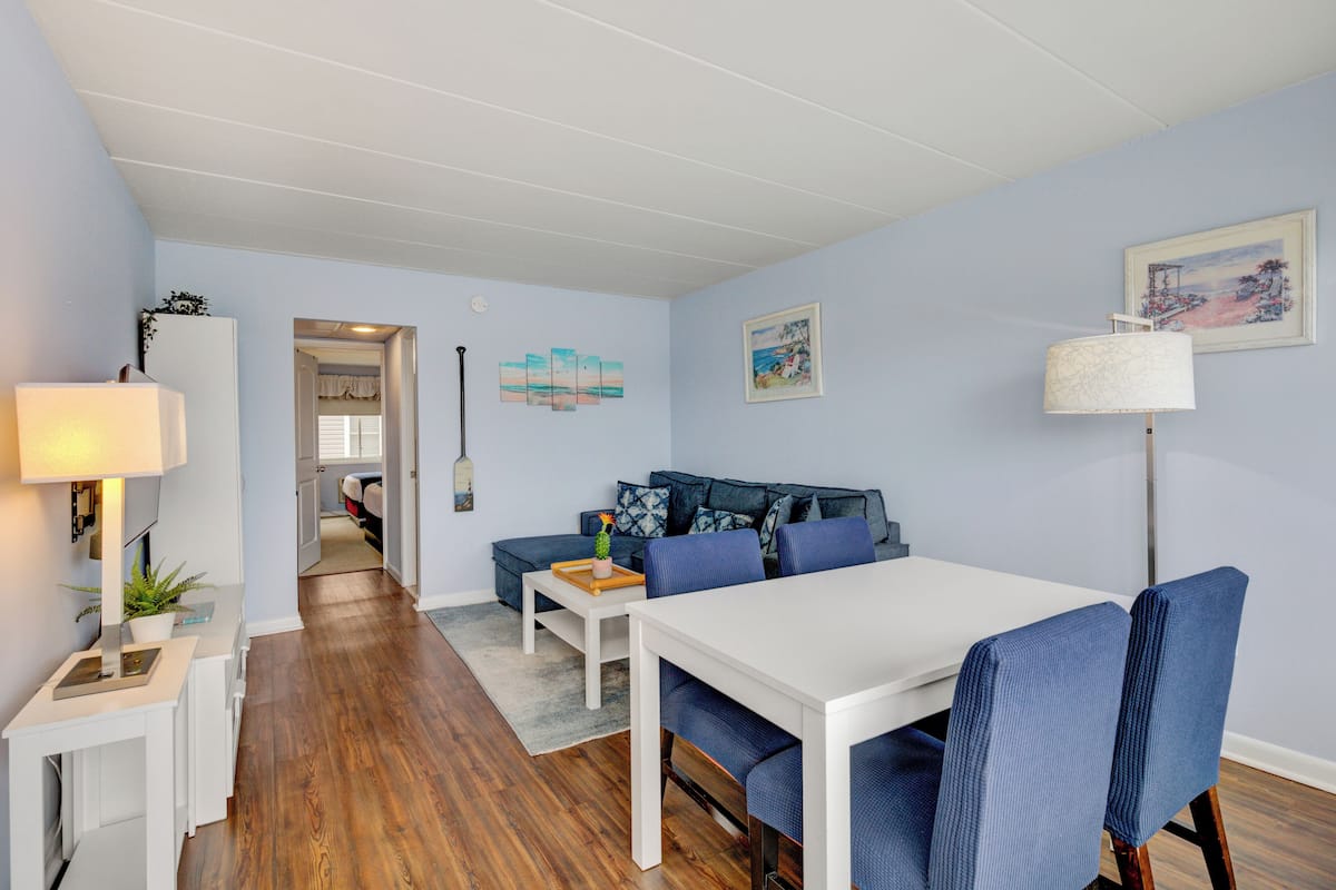 Una sala moderna con una mesa de comedor blanca, sillas tapizadas en azul, un sofá con cojines estampados y una lámpara de pie.
