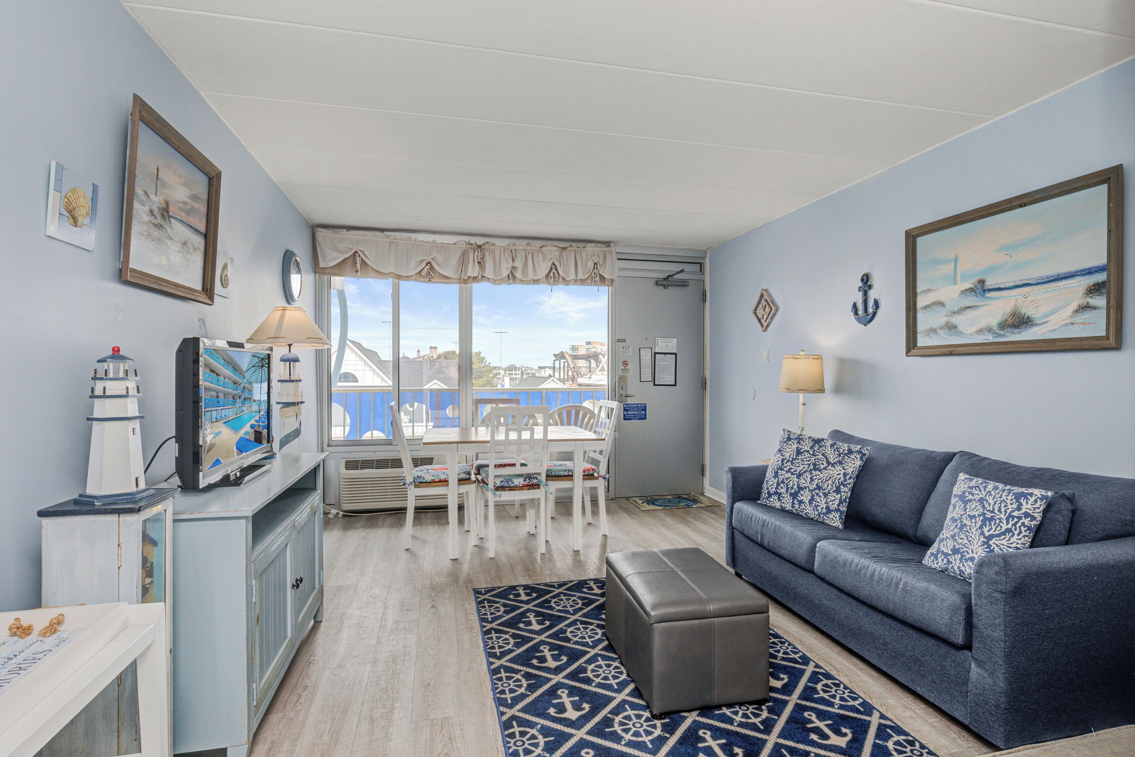 A living room with a blue sofa, a TV, a dining table with chairs, and a view of the city through large windows.