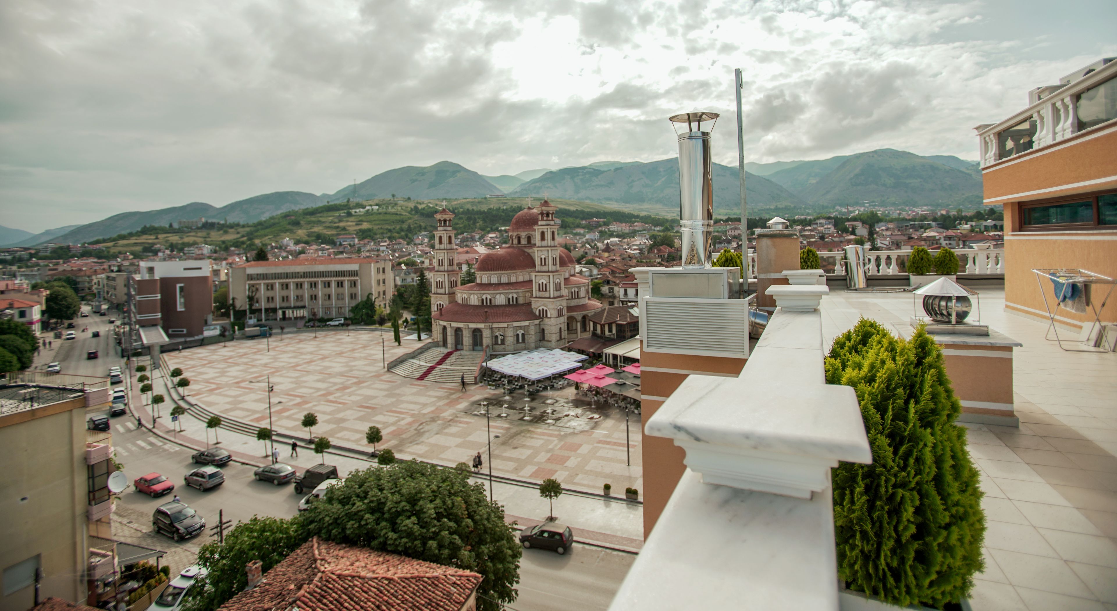 A plaza with a large building featuring a dome and bell tower, surrounded by modern buildings and a mountainous backdrop.