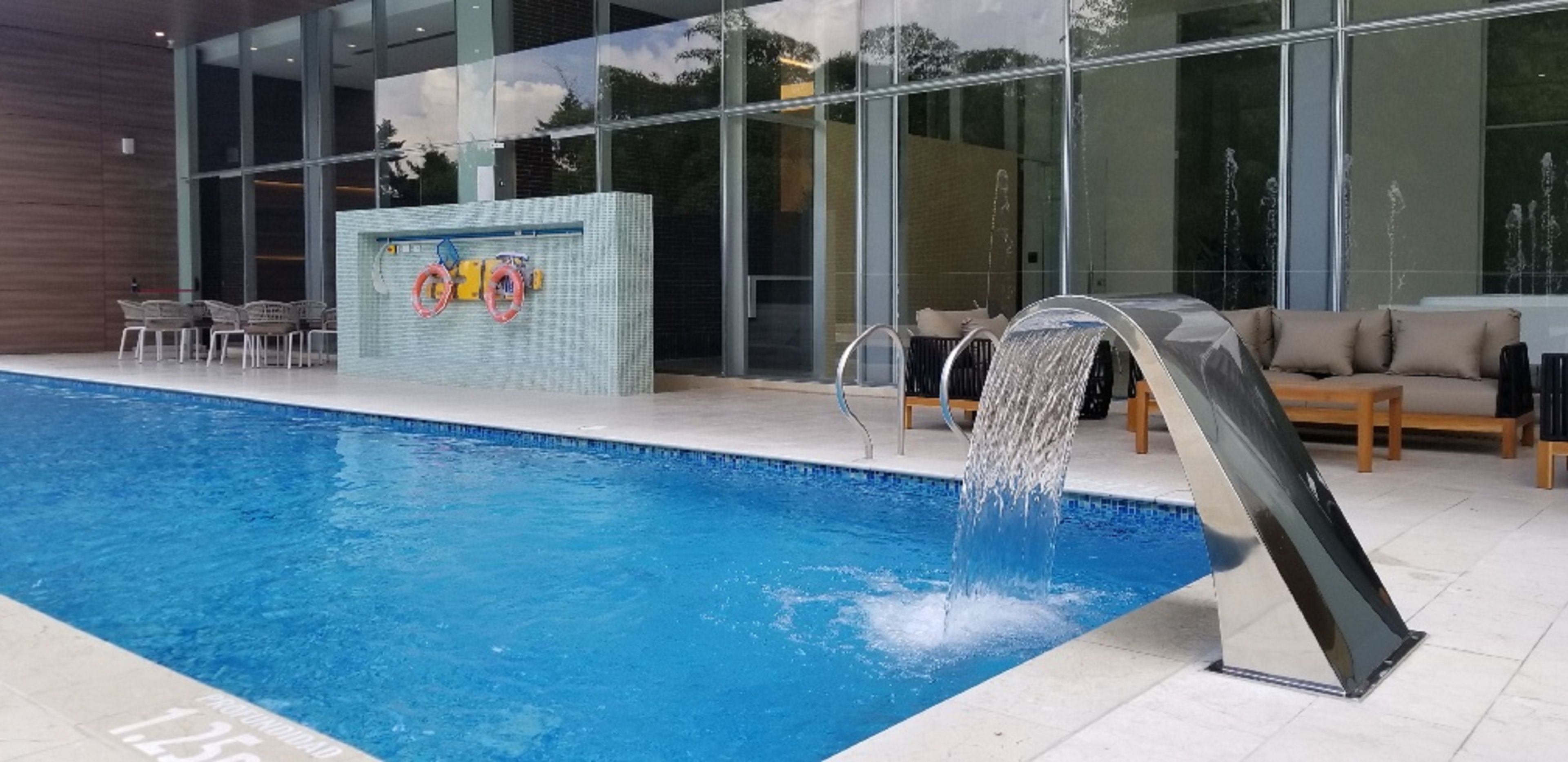 An indoor swimming pool with a water slide, lounge chairs, and a glass wall reflecting the sky.