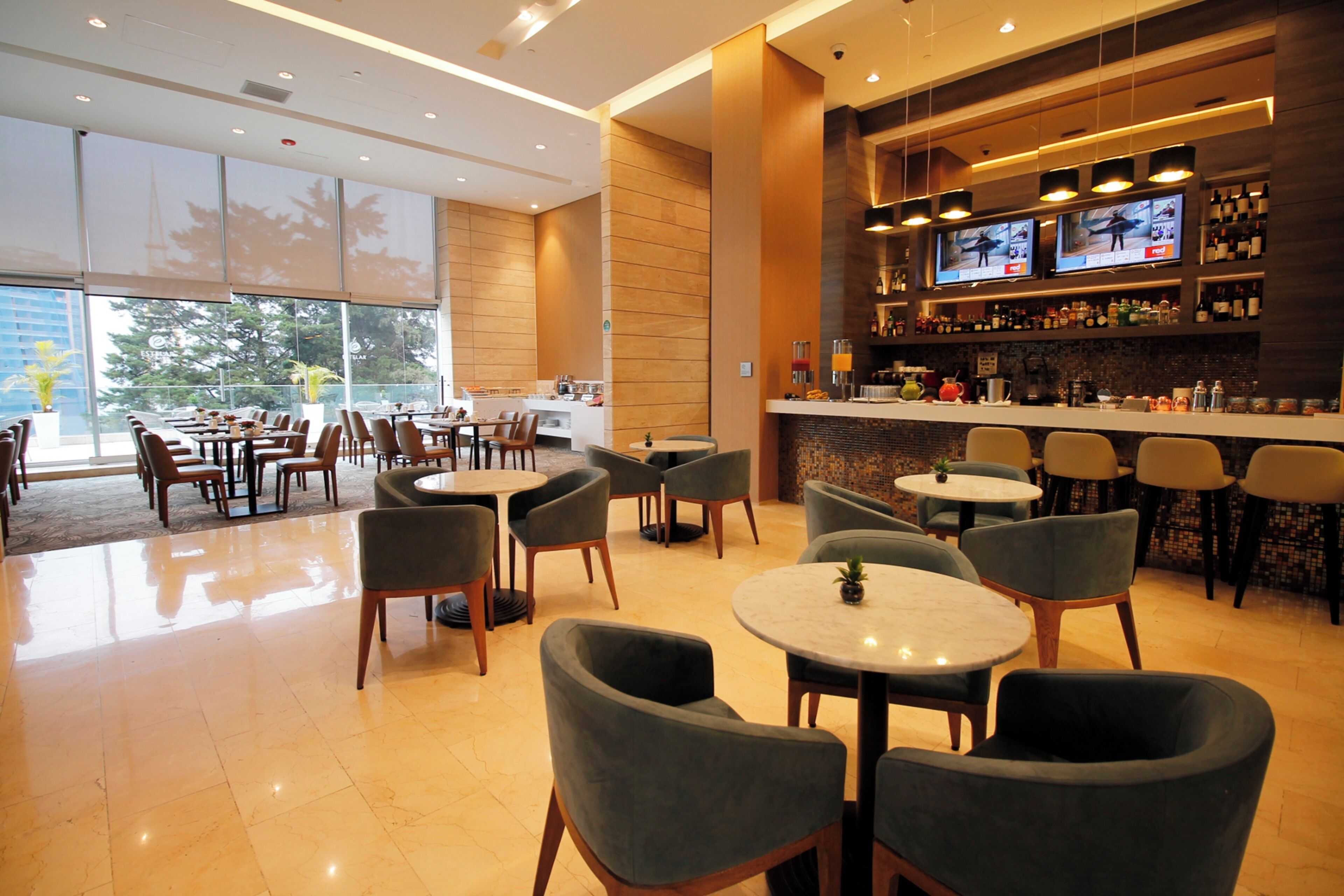 A hotel lobby with multiple seating areas, a bar with televisions, and a view of the outside.