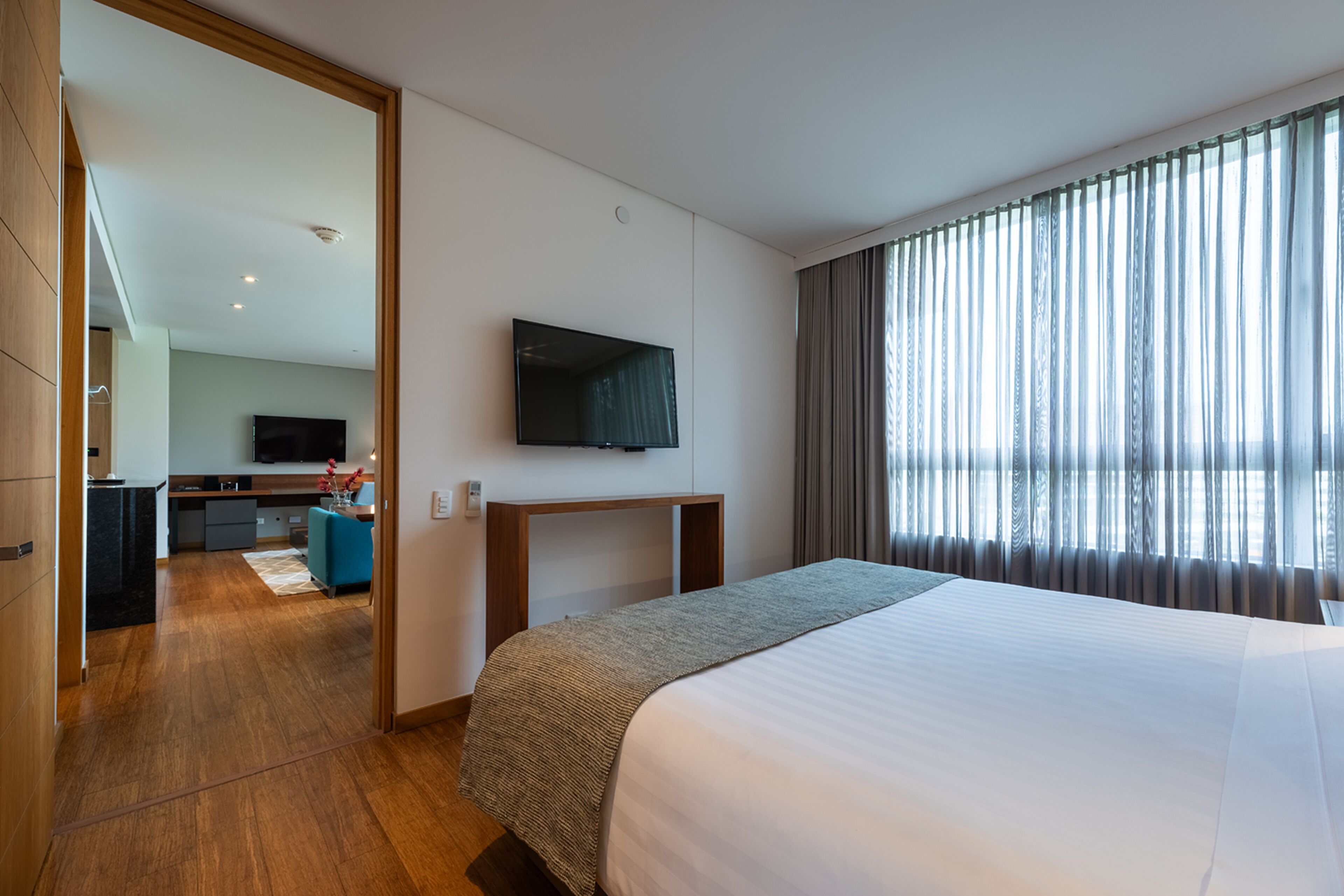 A modern hotel room with a large bed, a flat-screen TV mounted on the wall, a desk with a chair, and a view of the outdoors through sheer curtains.
