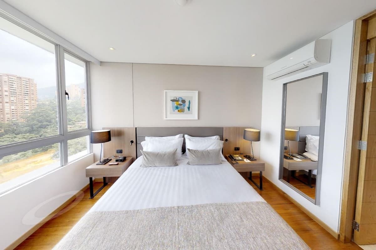 A modern hotel room with a large bed, two bedside tables with lamps, a large window, and a mirror.