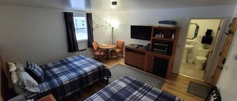 Habitación de hotel pequeña con dos camas, un escritorio, una silla, un televisor y un baño visible a través de una puerta abierta.