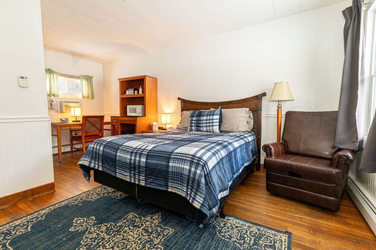 A bedroom with a bed, a brown leather chair, a wooden nightstand, a lamp, and a small desk with a lamp.