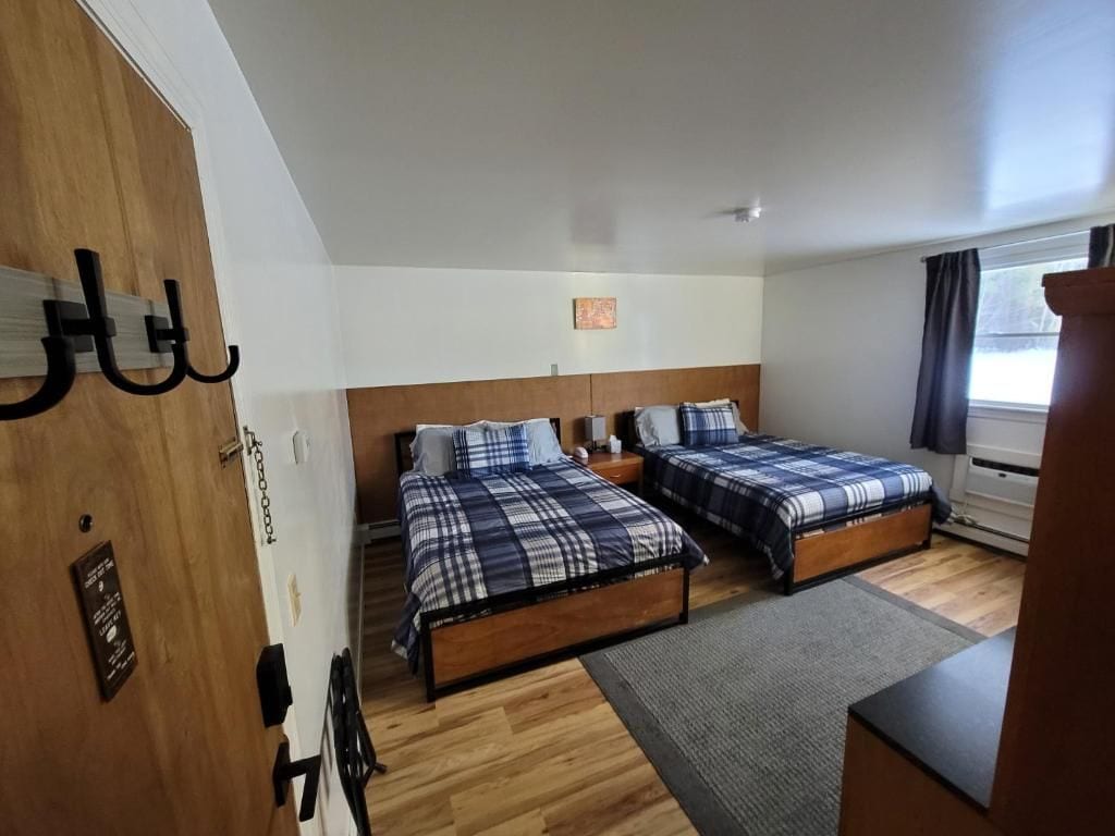 A room with two beds, a wooden floor, a small rug, a nightstand, and a window with curtains.
