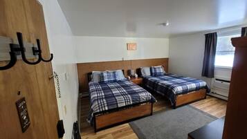 A room with two beds, a wooden floor, a small rug, a nightstand, and a window with curtains.