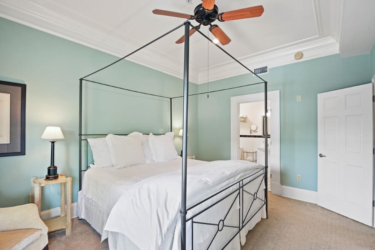 A four-poster canopy bed with a metal frame, a ceiling fan, a bedside table with a lamp, and a door to a bathroom.