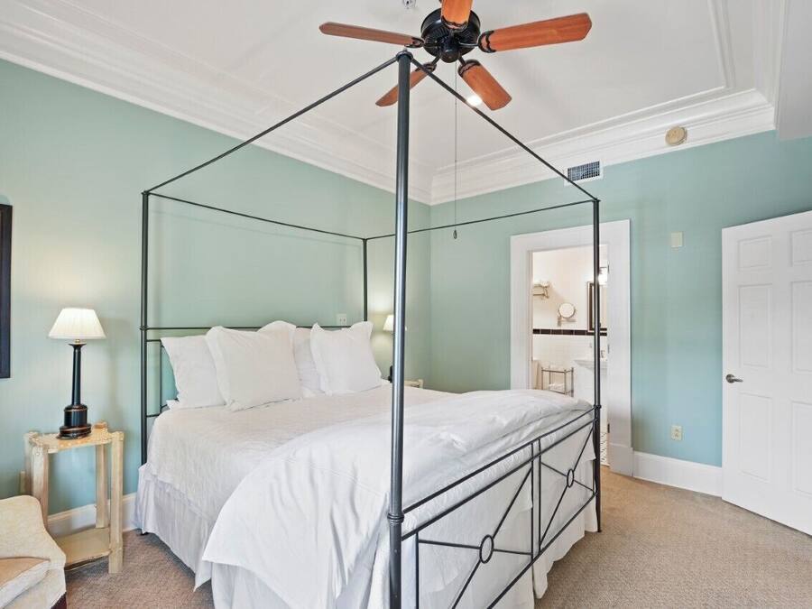 A four-poster canopy bed with a metal frame, a ceiling fan, a bedside table with a lamp, and a door to a bathroom.