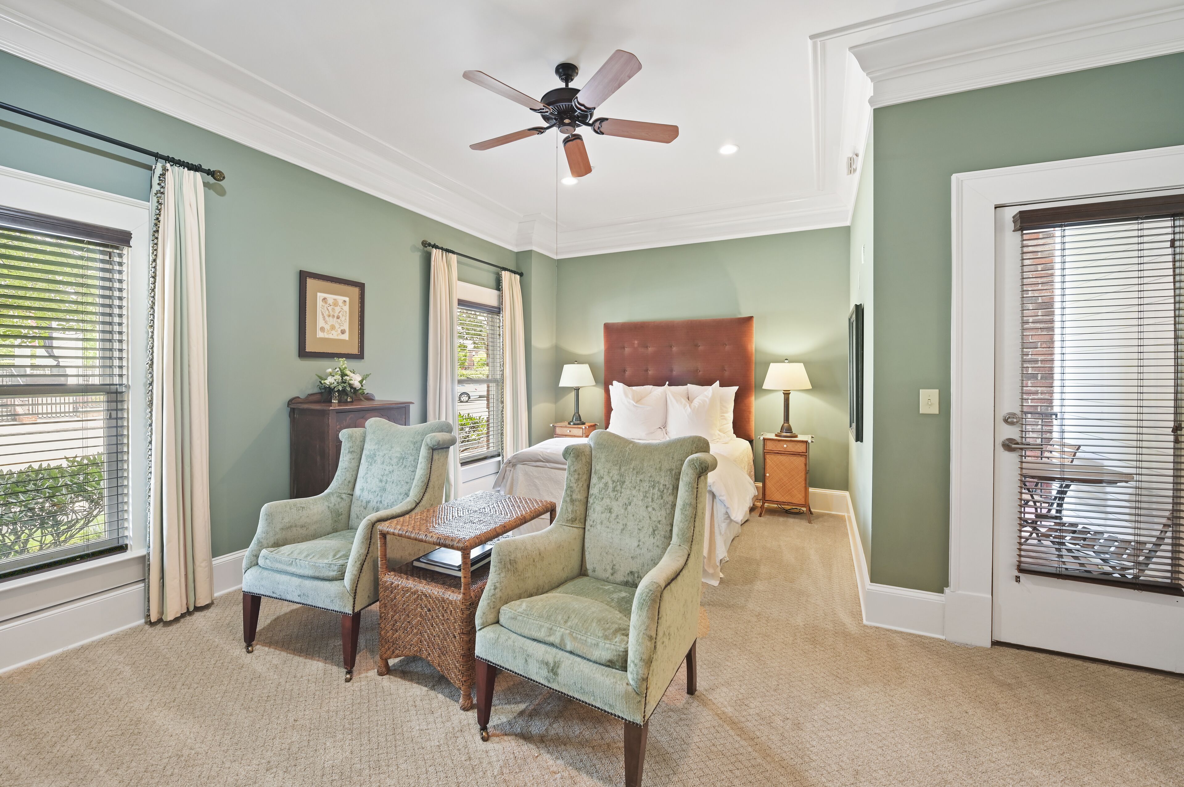 A bedroom with a large bed, two armchairs, a small table, a ceiling fan, and a view of the outdoors through the window.