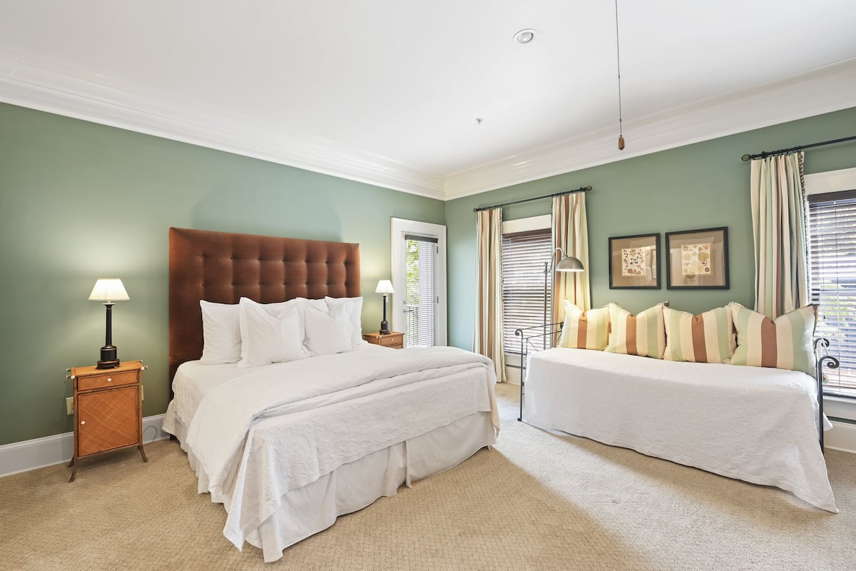 A bedroom with a large bed, a smaller bed, a nightstand, a lamp, a window with curtains, and a door.