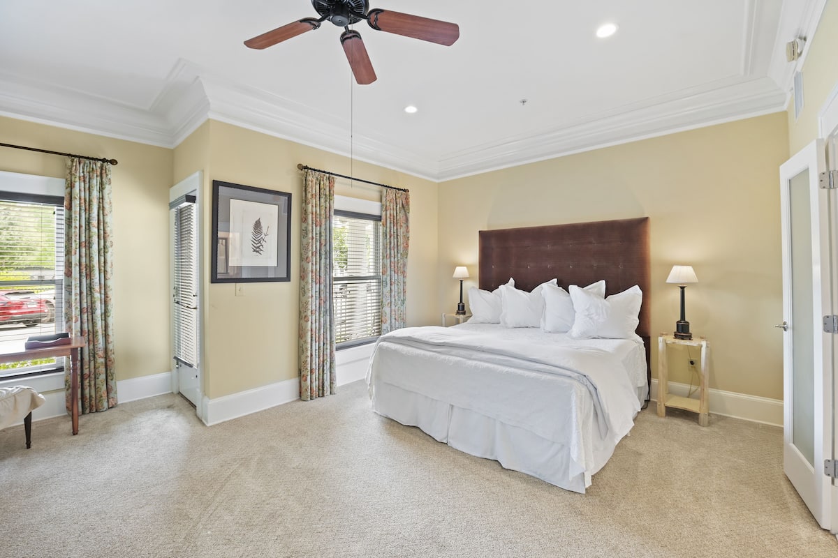 A bedroom with a large bed, two bedside tables with lamps, a ceiling fan, and a window with curtains.