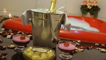 Una botella de champán en un balde de metal, dos copas pequeñas con bebidas rosadas, y una bandeja con objetos amarillos sobre una encimera adornada con pétalos de rosa.