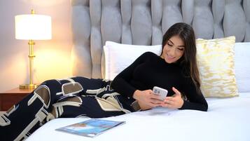 Una persona recostada en una cama con un teléfono inteligente, una lámpara y un libro sobre la mesita de noche.