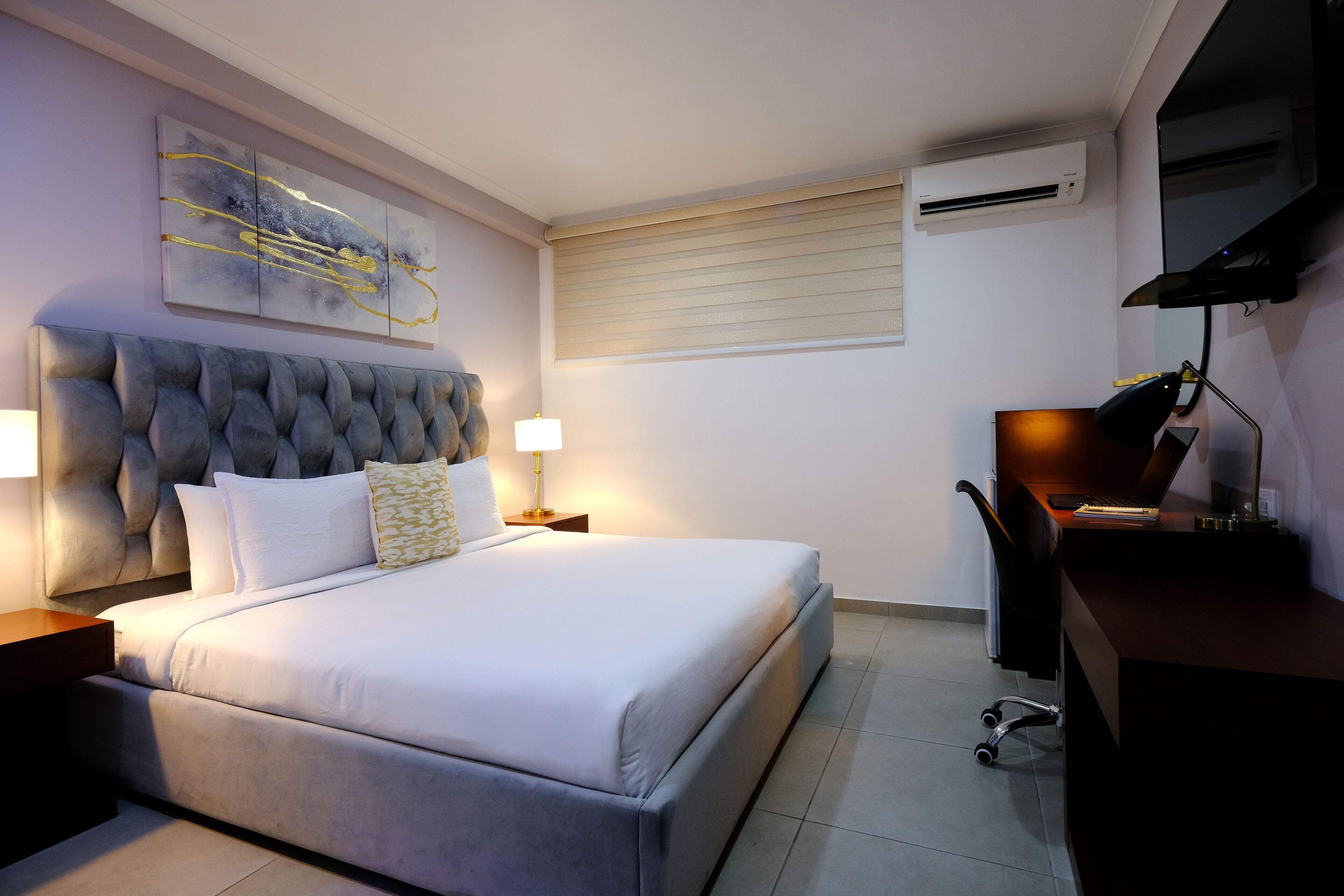 A modern bedroom with a tufted headboard, a large bed, bedside lamps, a desk with a chair, and a mounted air conditioner.