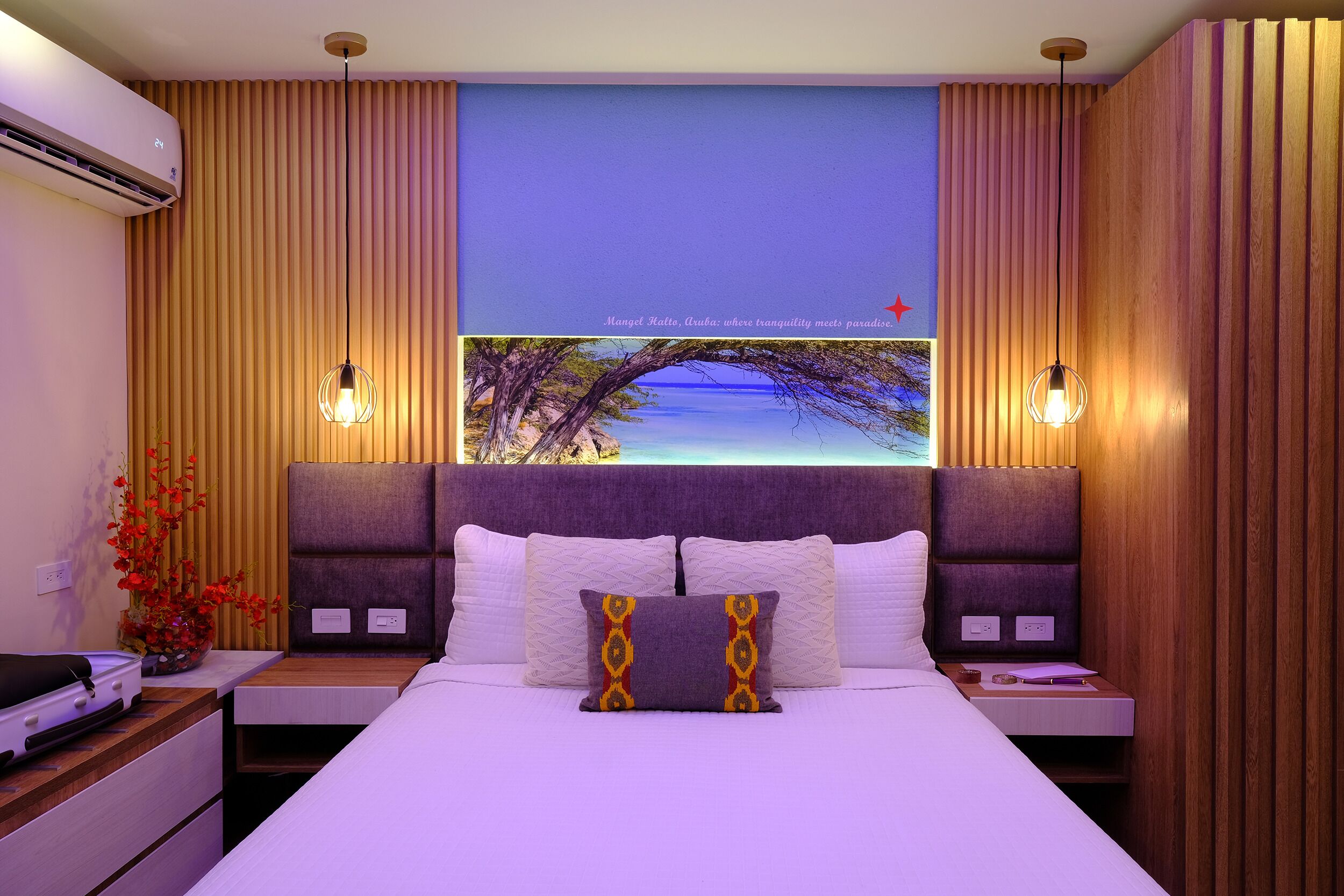 A hotel room with a large bed, two bedside tables, a nightstand, a wall-mounted air conditioner, and a decorative wall with a scenic view.