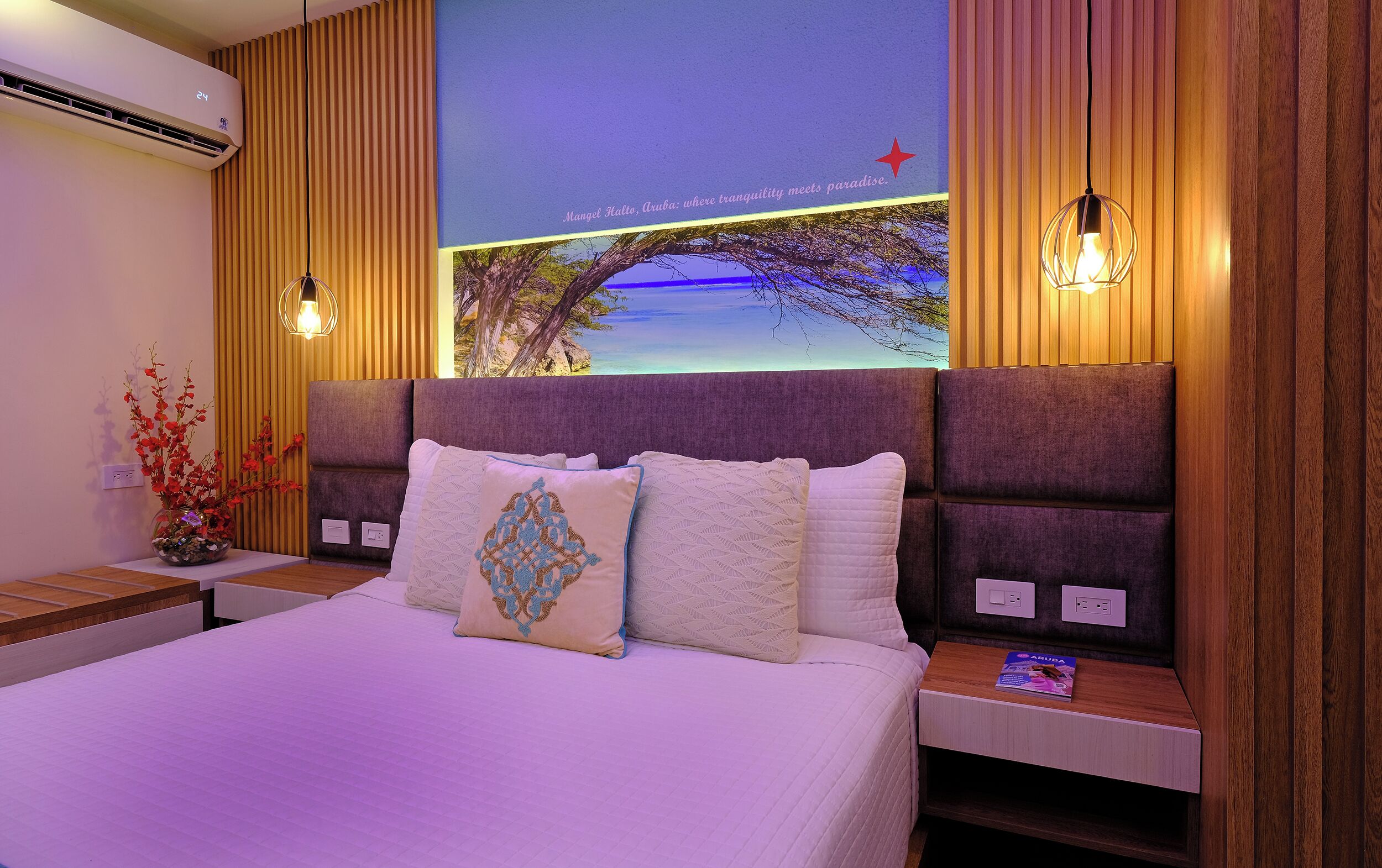 Hotelkamer met een groot bed, een nachtkastje met lamp, een wandairconditioner en een muurschildering van een brug en bomen.