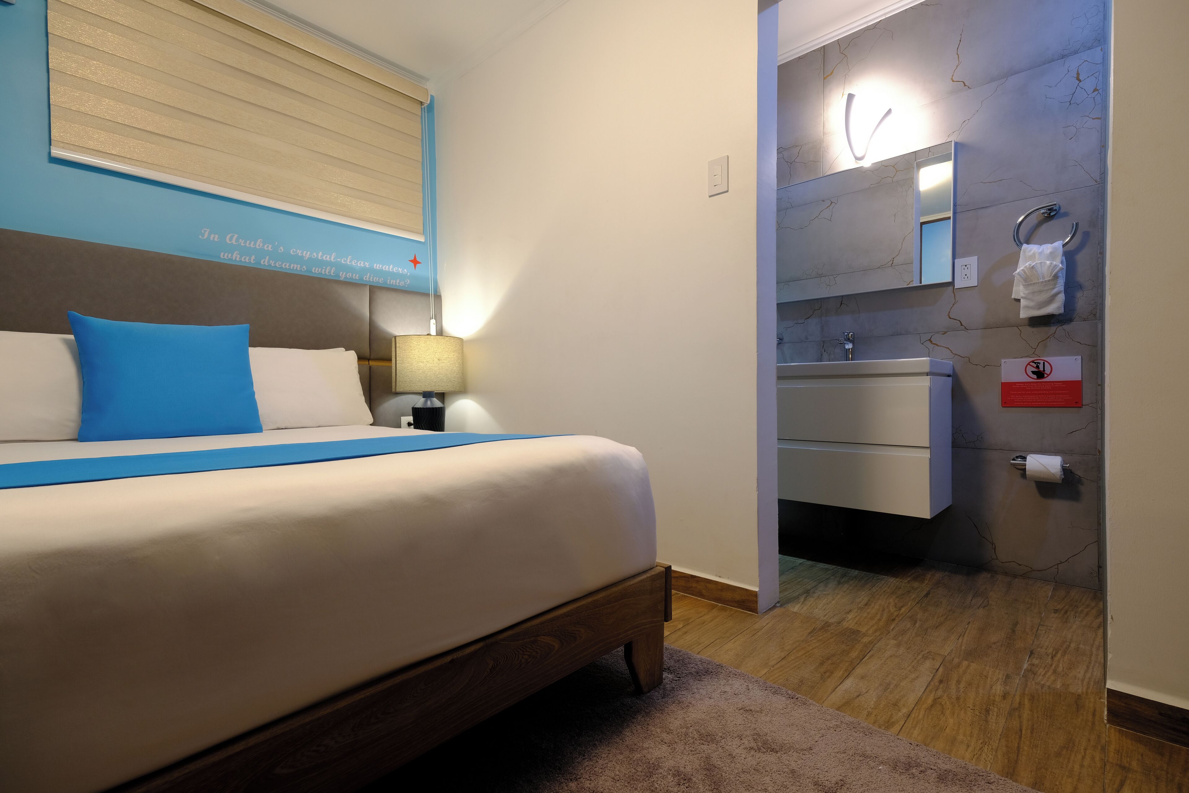 Un dormitorio con cama, mesita de noche, lámpara, baño con lavabo y espejo, y un cartel con un símbolo de prohibido.