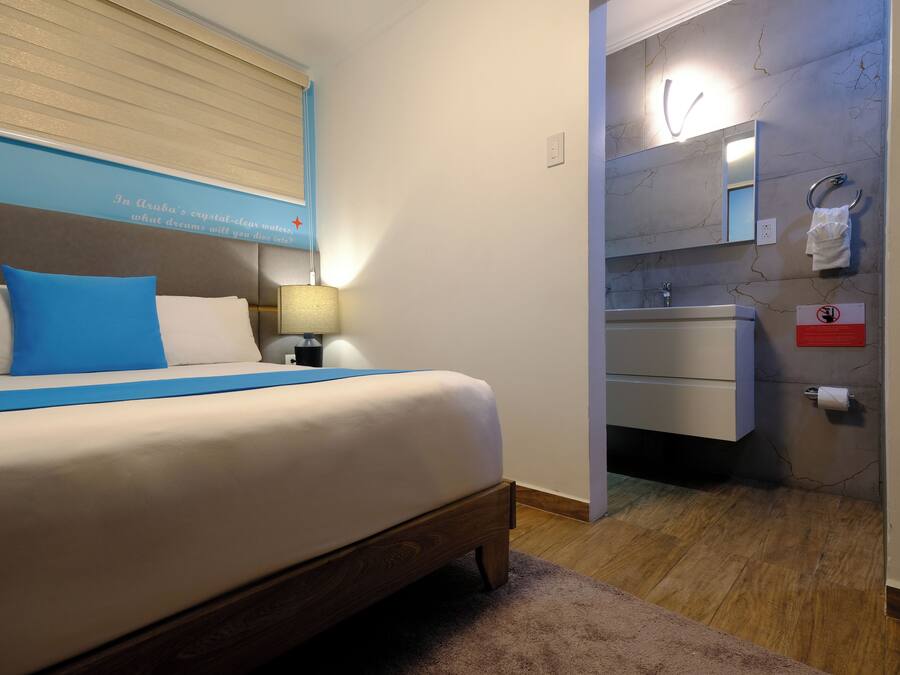 Un dormitorio con cama, mesita de noche, lámpara, baño con lavabo y espejo, y un cartel con un símbolo de prohibido.