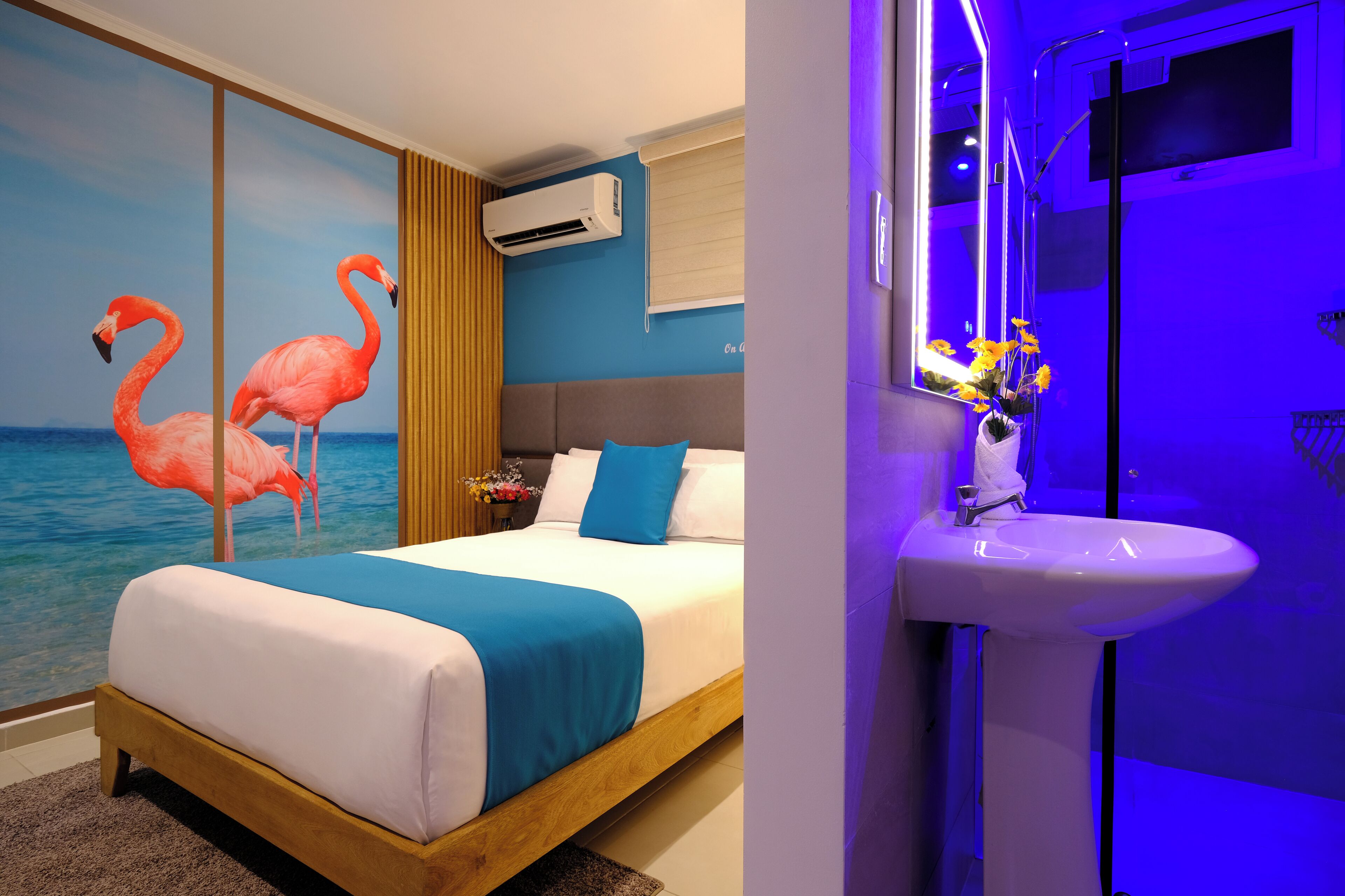 A hotel room with a bed, a wall-mounted air conditioner, a bathroom with a sink and mirror, and a wall with a painting of flamingos.