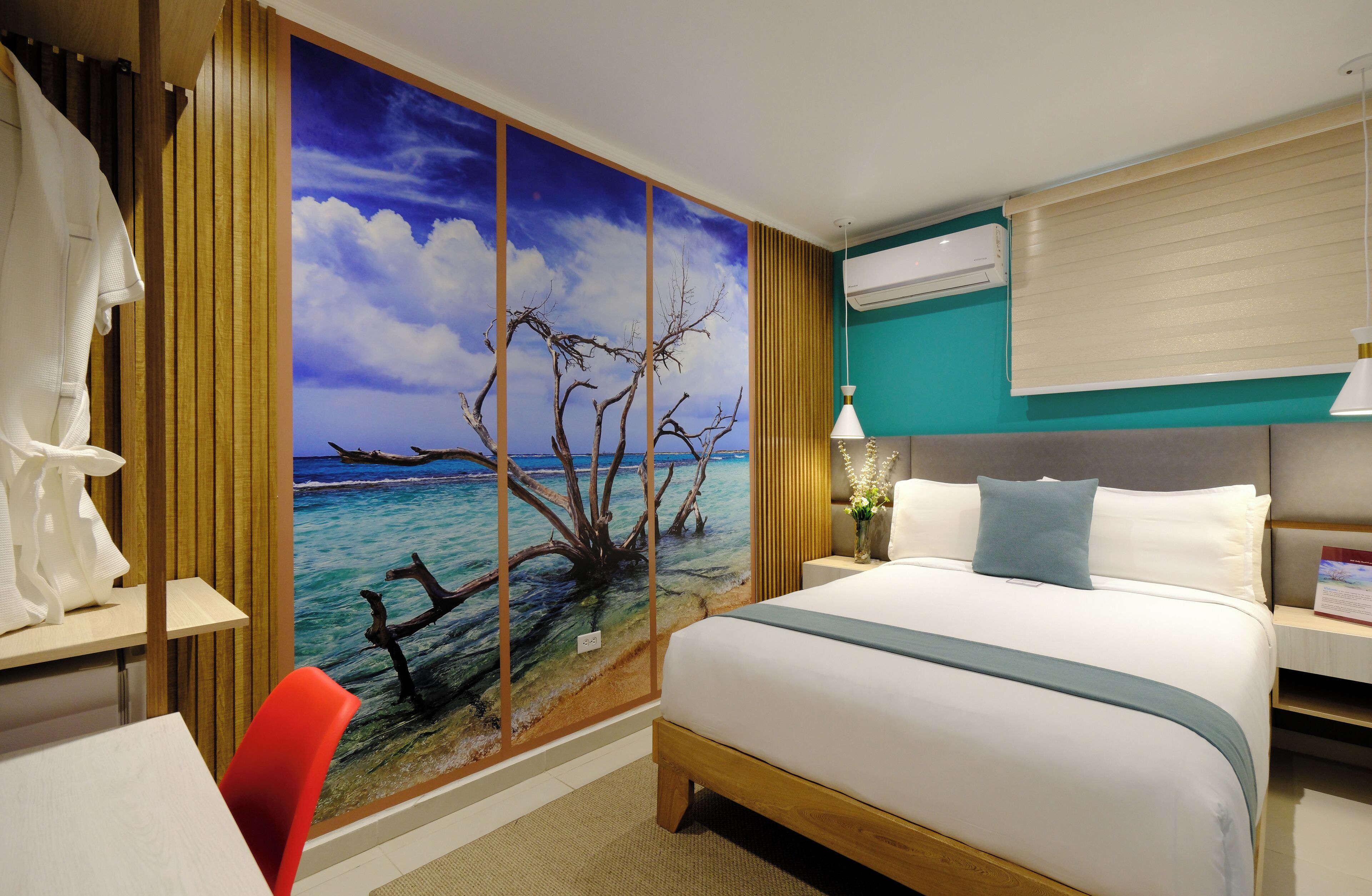 Habitación de hotel con una cama grande, un escritorio, una silla, un ventanal con vista al mar y un mural en la pared que representa un árbol y el cielo.